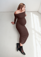 Woman wearing a brown off-shoulder dress against a white wall.