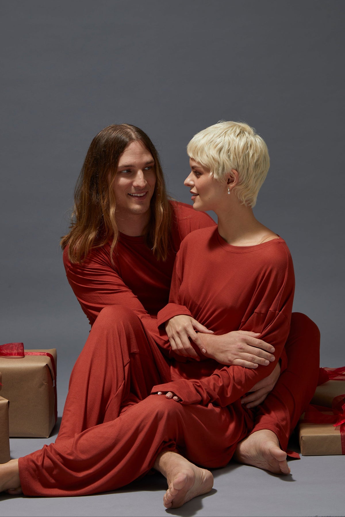 Two people in red lounge outfits sitting together with wrapped gifts on a gray background
