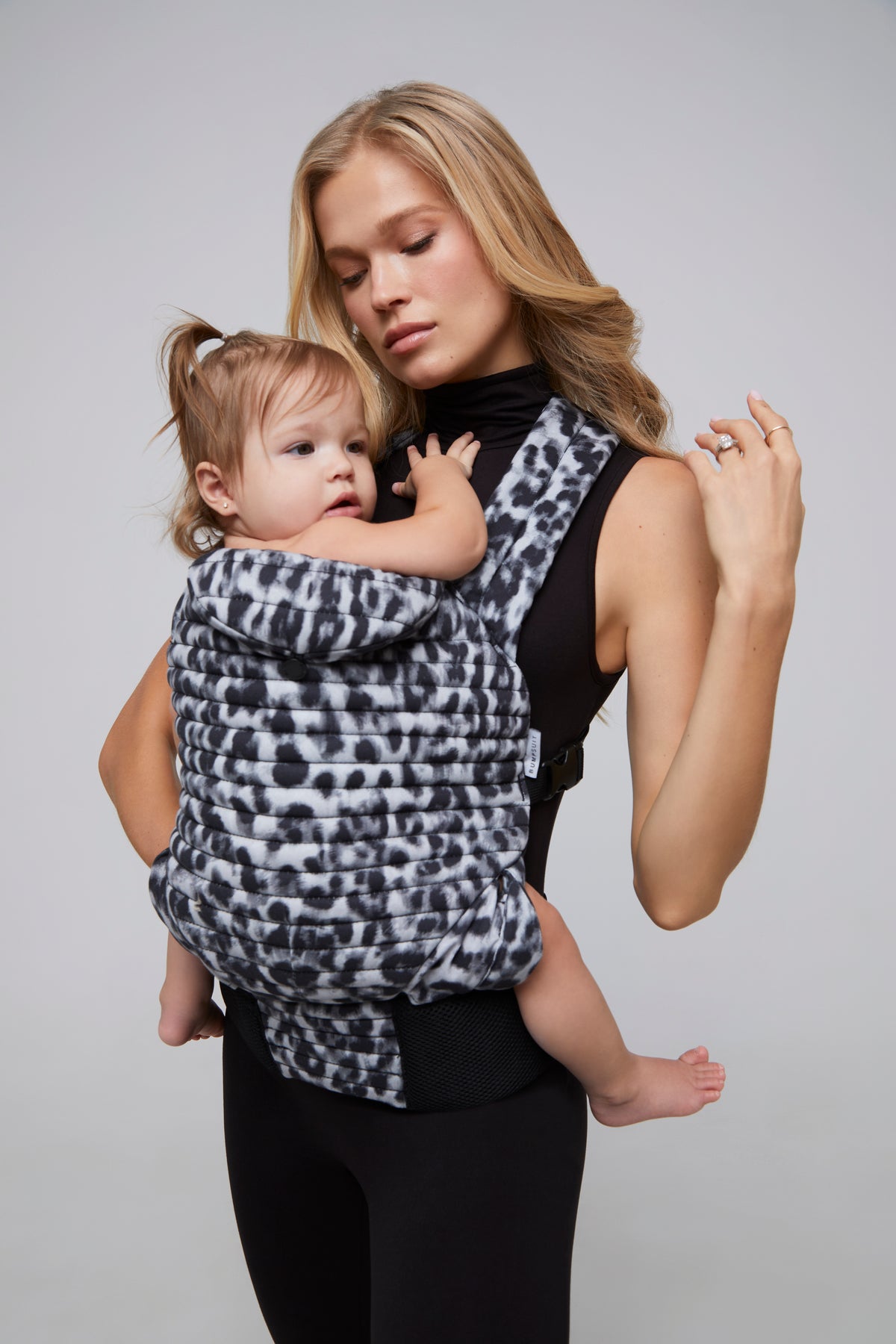 Woman holding a child in a monochrome leopard print baby carrier against a plain background