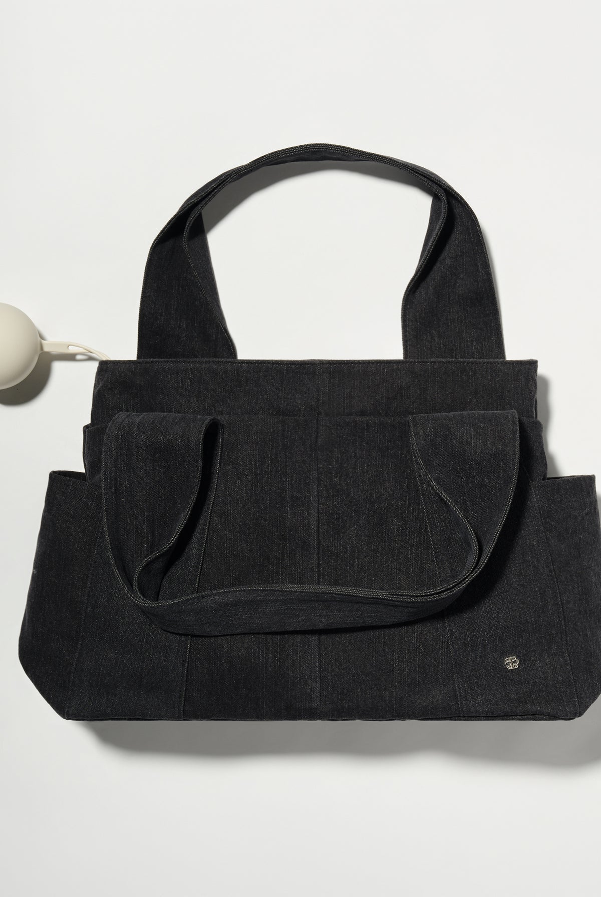 A black denim fabric diaper bag with dual top handles and front slip pockets, shown on a plain light background with a small pacifier holder on the left.