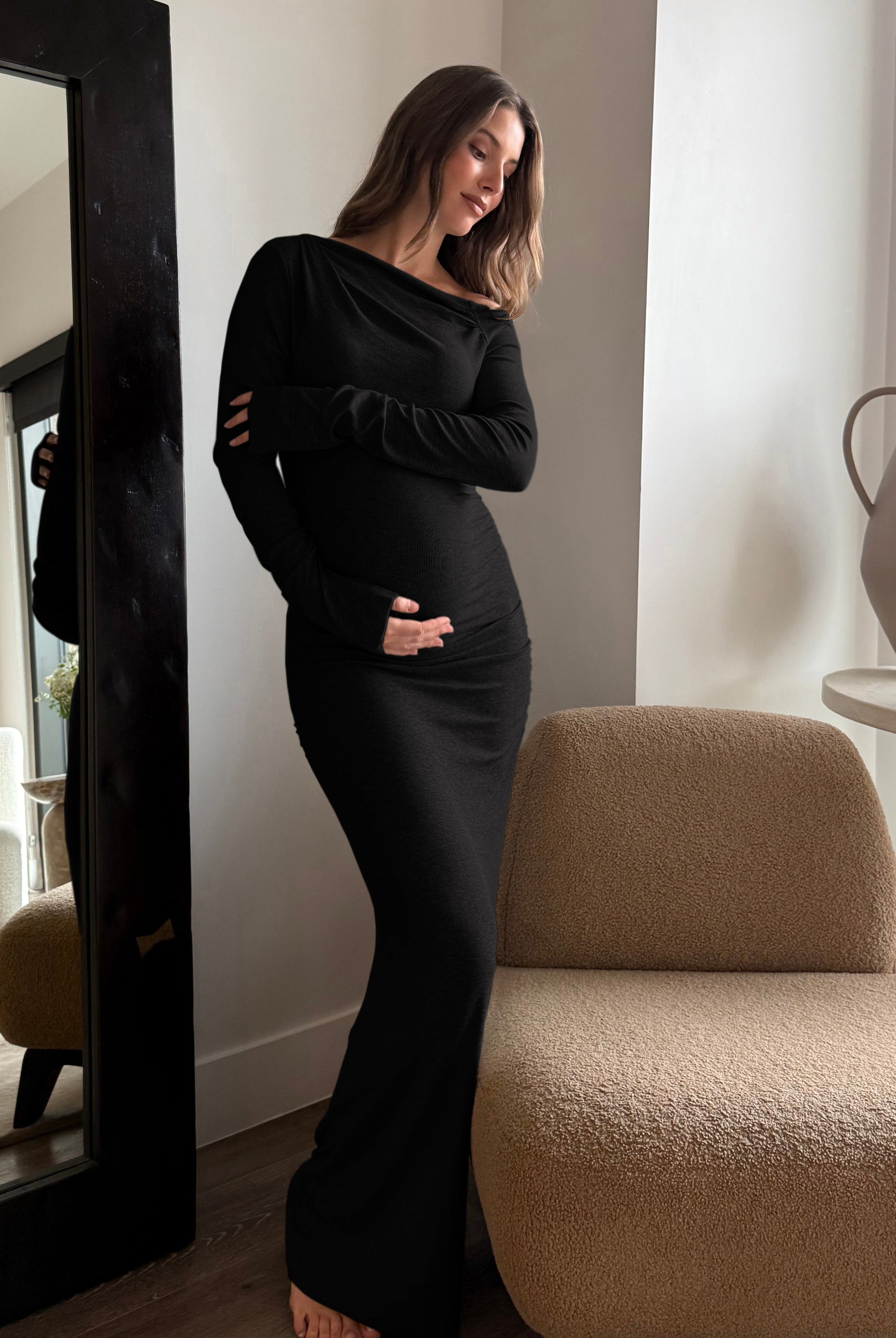 A pregnant woman wearing a Bumpsuit long-sleeve, off-the-shoulder, soft rib maternity dress in black, standing in a modern, sunlit room next to a large black-framed mirror.