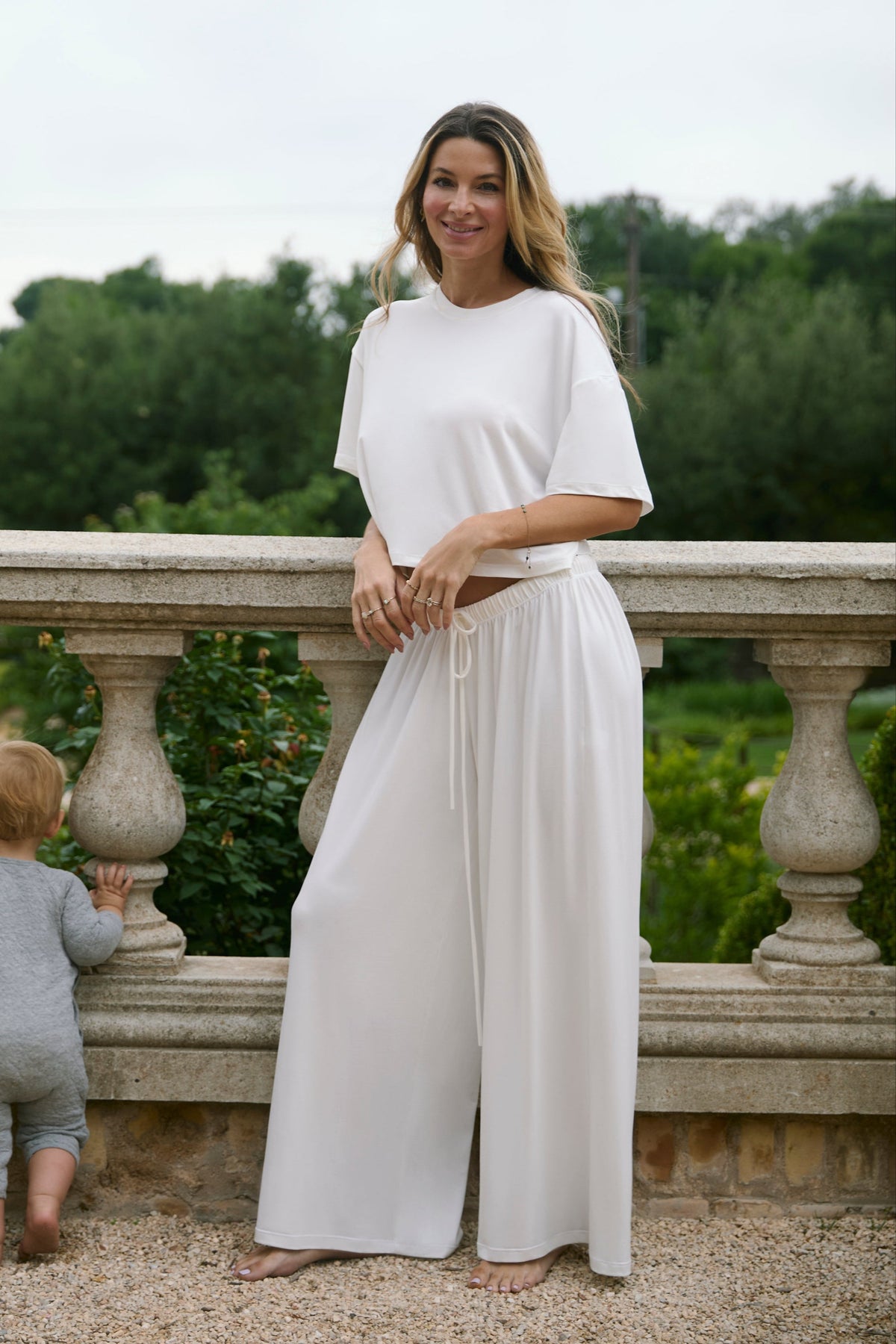 Woman wearing the Bumpsuit Maternity Cloud Short Sleeve Crop Tee and pants in Ivory with a toddler standing in an outdoor setting
