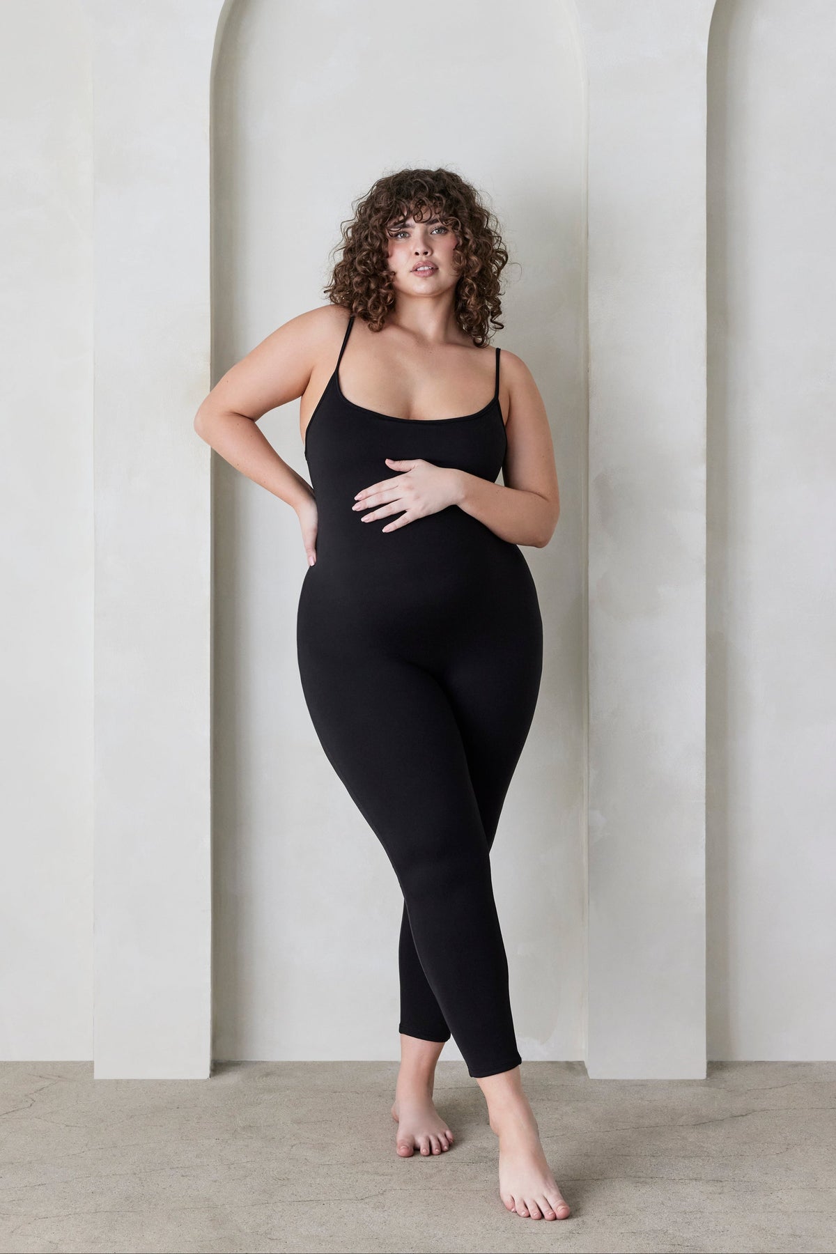 Woman wearing a black Bumpsuit sleeveless jumpsuit called the Kate standing in a minimalistic setting.