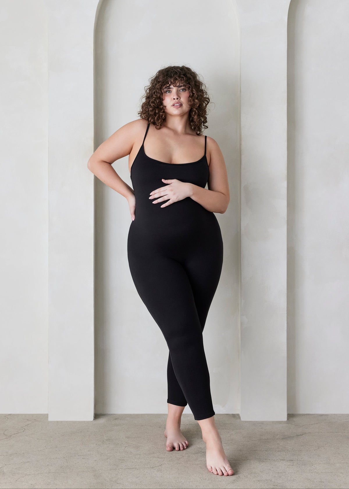 Woman wearing a black Bumpsuit sleeveless jumpsuit called the Kate standing in a minimalistic setting.