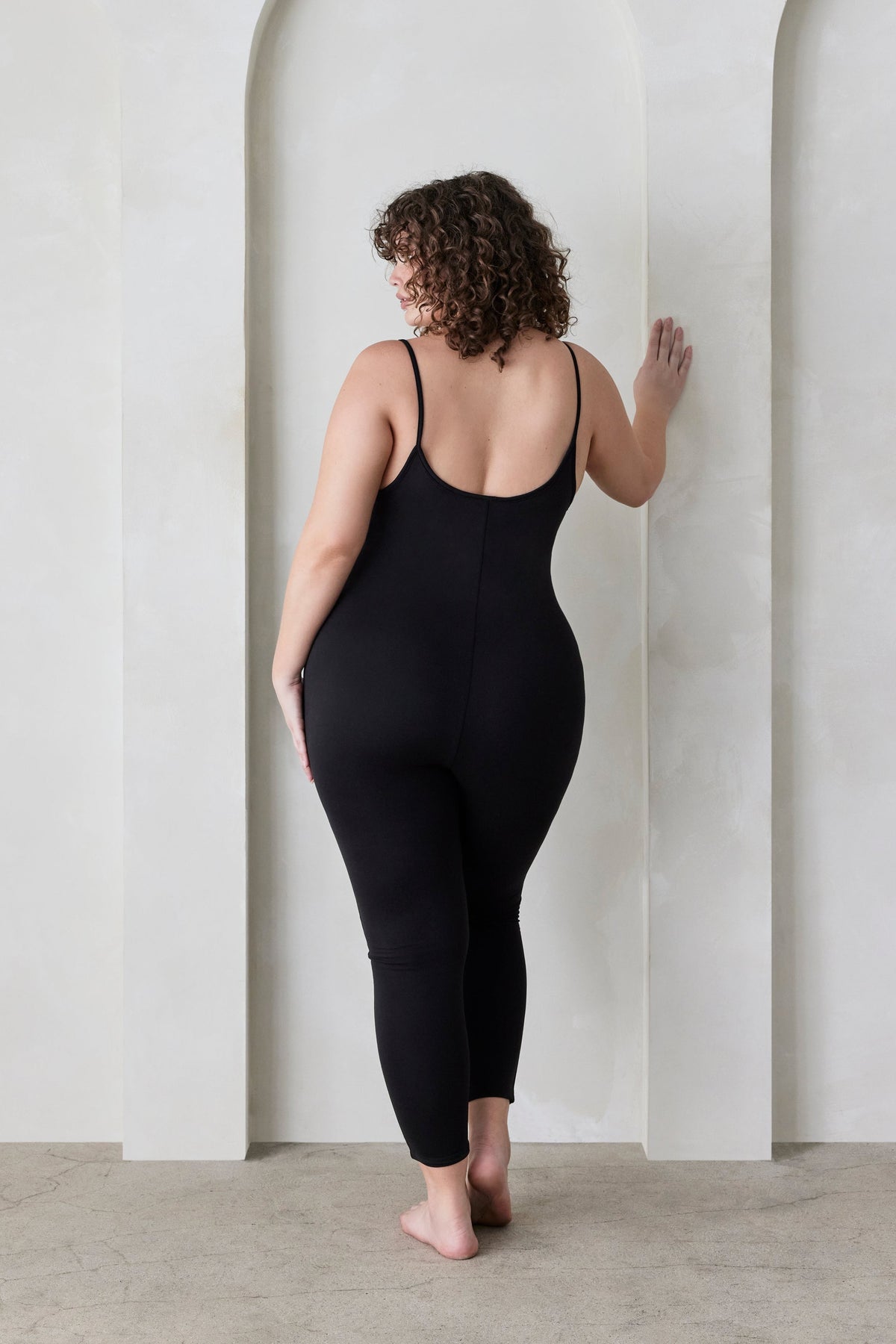 Back view of a woman wearing a Bumpsuit black sleeveless jumpsuit called the Kate standing in a minimalistic setting.