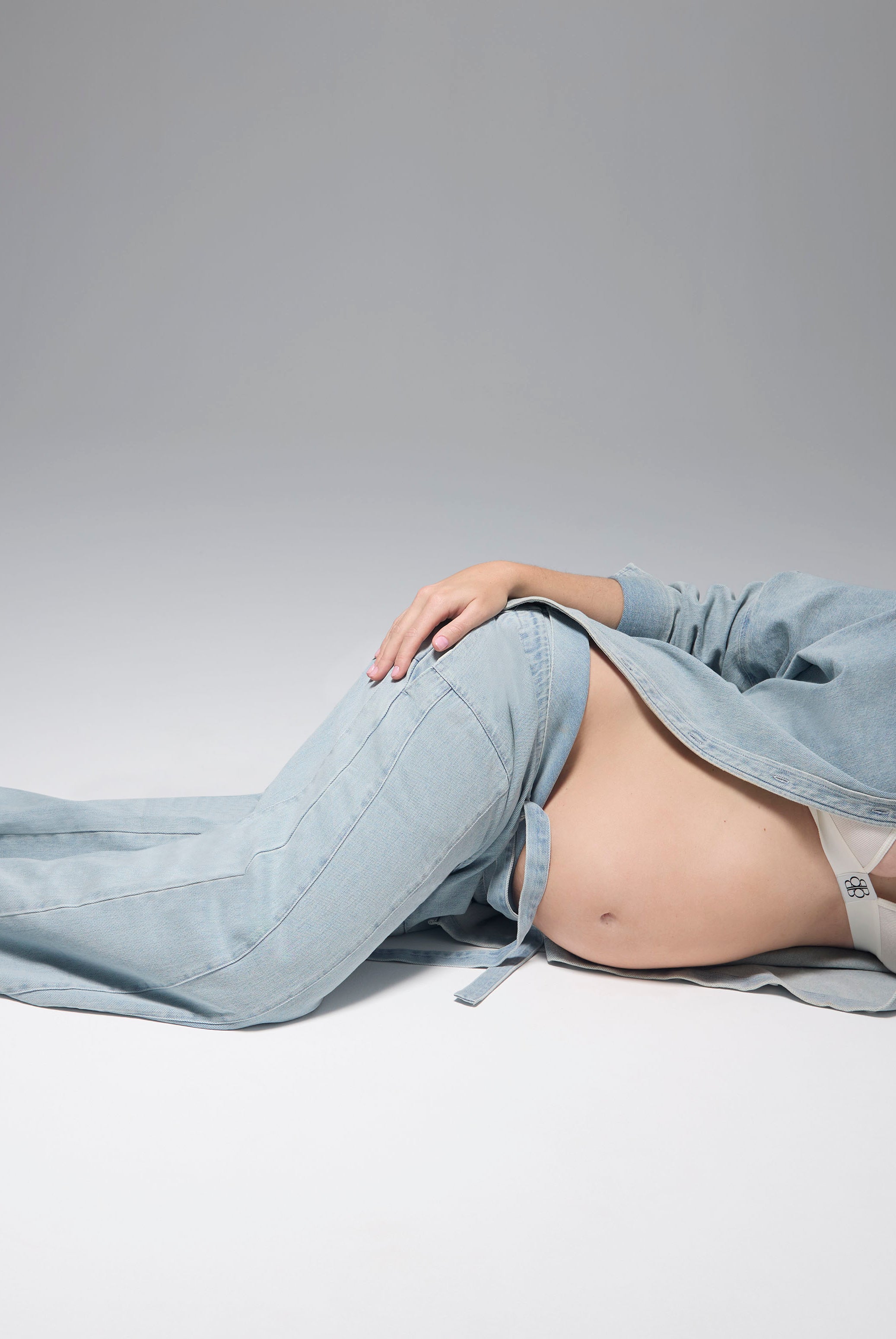 Pregnant woman lying down wearing Bumpsuits light blue denim button up shirt and jeans against a gray background