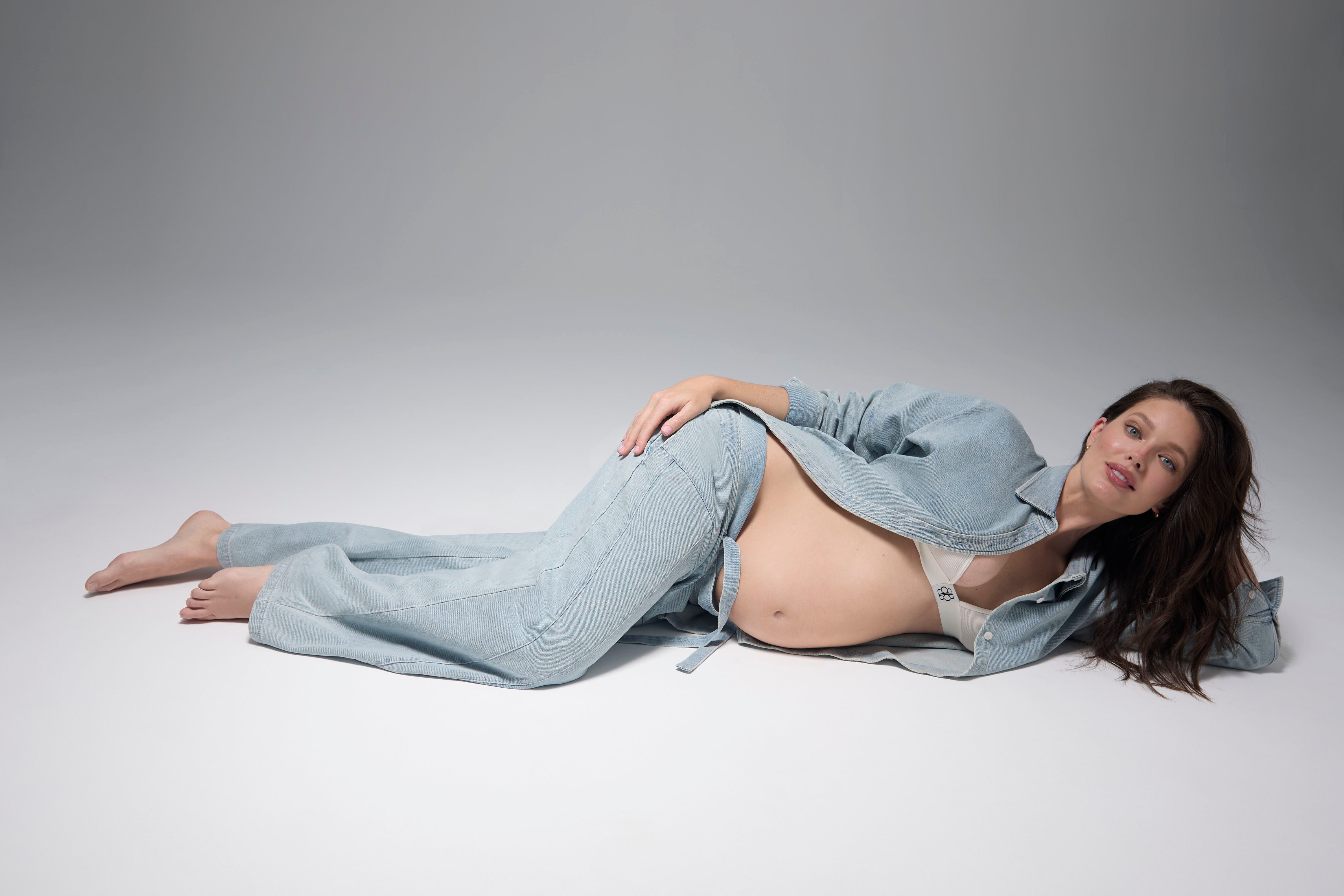 Pregnant woman lying down wearing Bumpsuits light blue denim button up shirt and jeans against a gray background