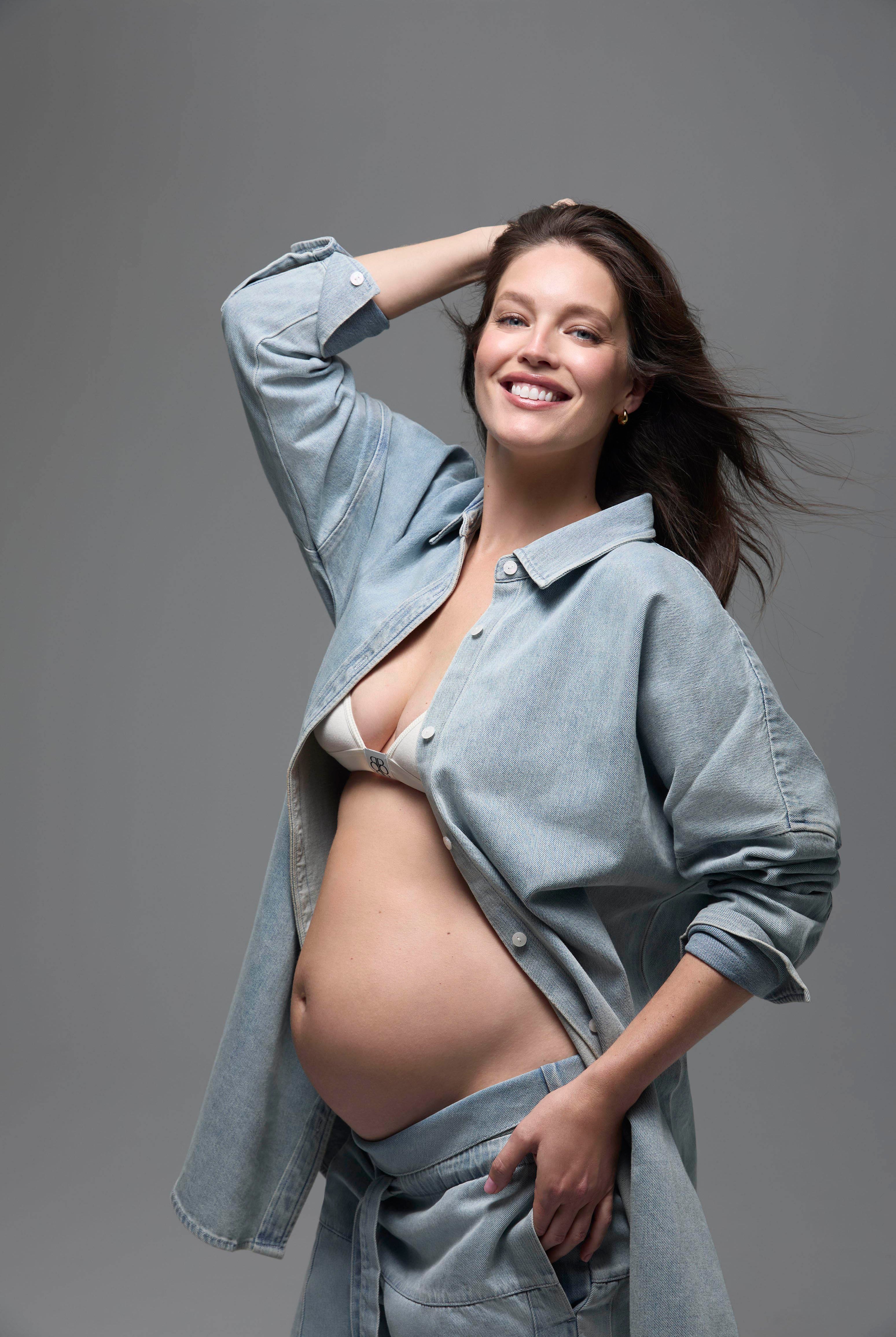Pregnant woman wearing a light blue denim shirt and pants with a white cotton bra against a gray background