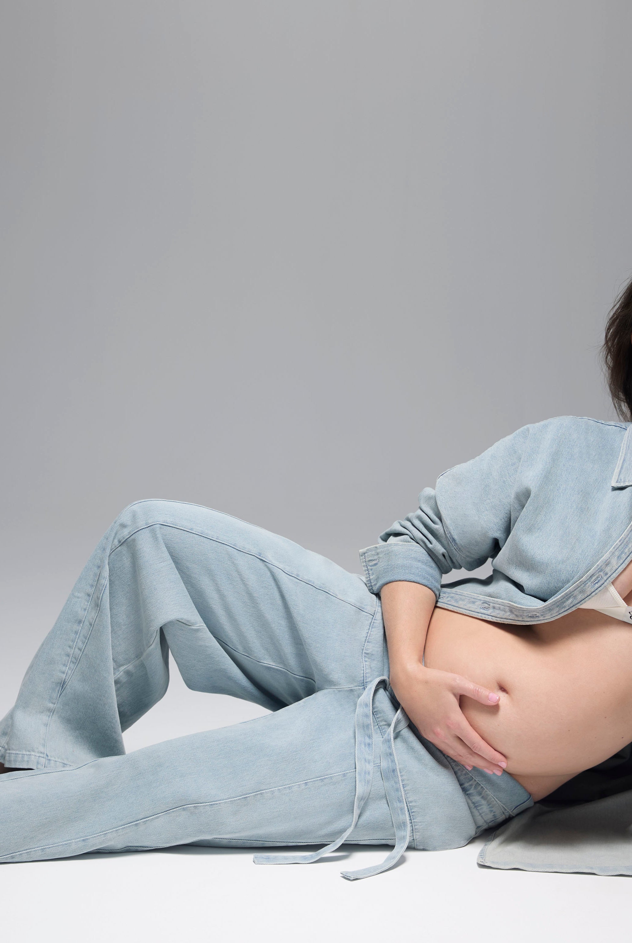 Pregnant woman lying on a gray background wearing light blue denim pants and a denim jacket with a white cotton bra