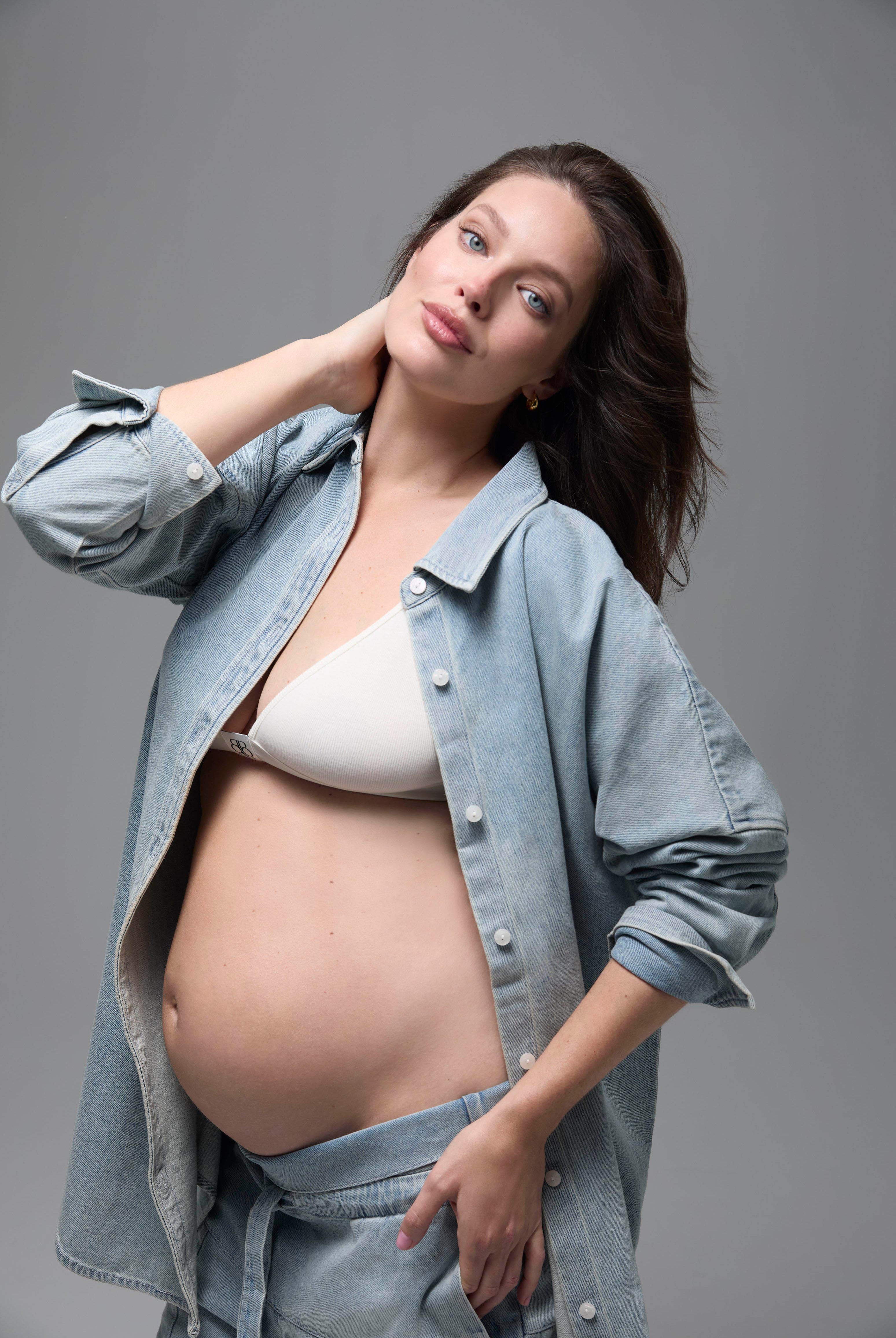Pregnant woman wearing a light blue denim shirt over a white bra on a gray background