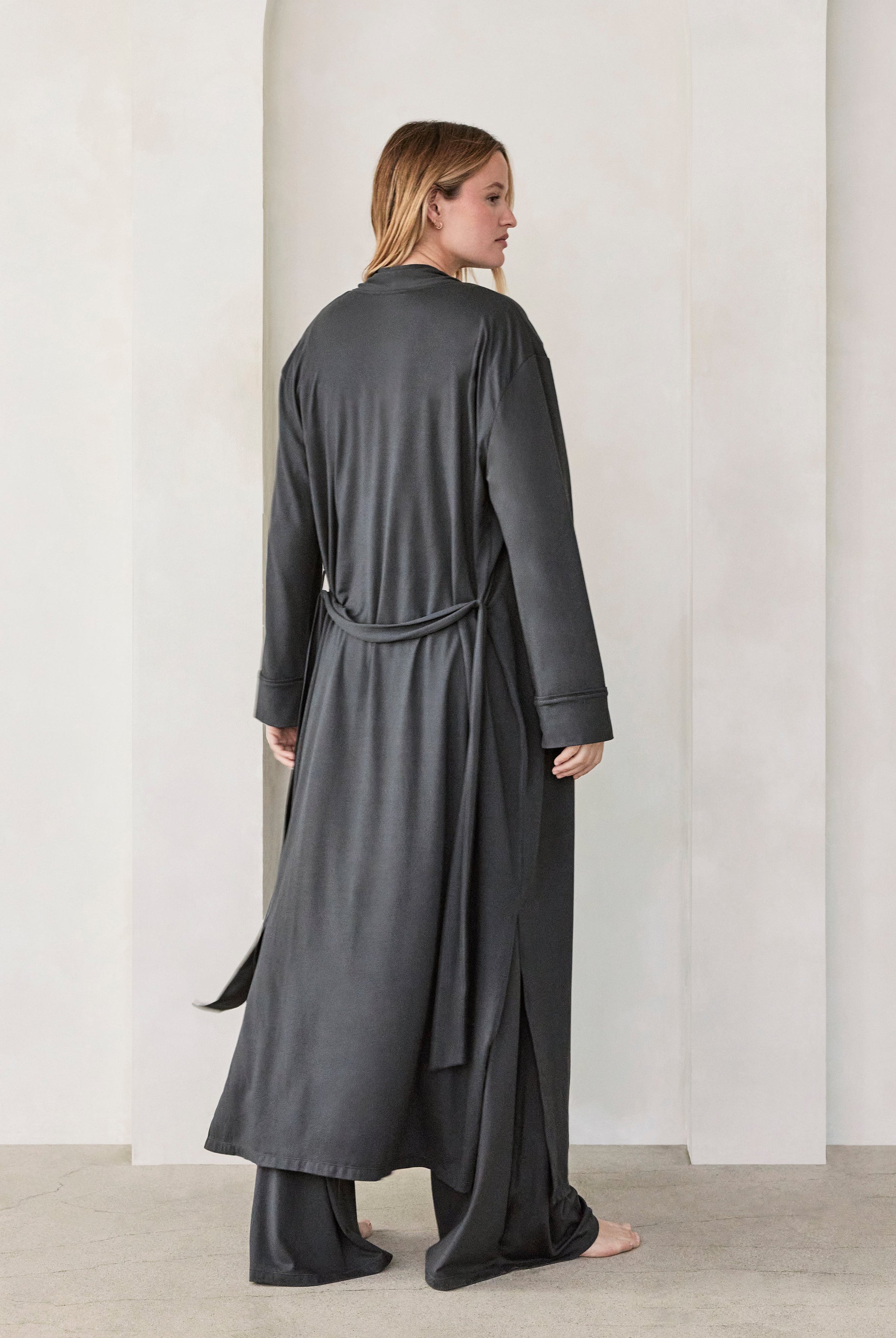 Woman wearing a dark grey slate Bumpsuit Cloud outfit standing in a minimalistic room with beige walls and arches.