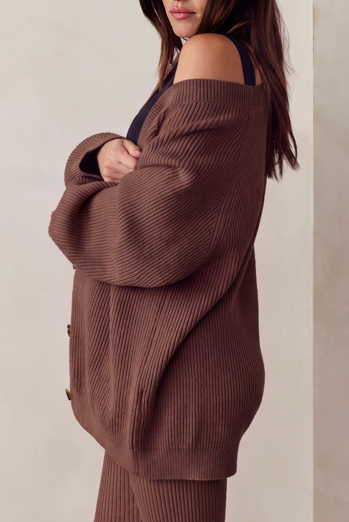 A pregnant woman wearing a matching brown ribbed knit cardigan and leggings set, standing against a neutral beige wall.