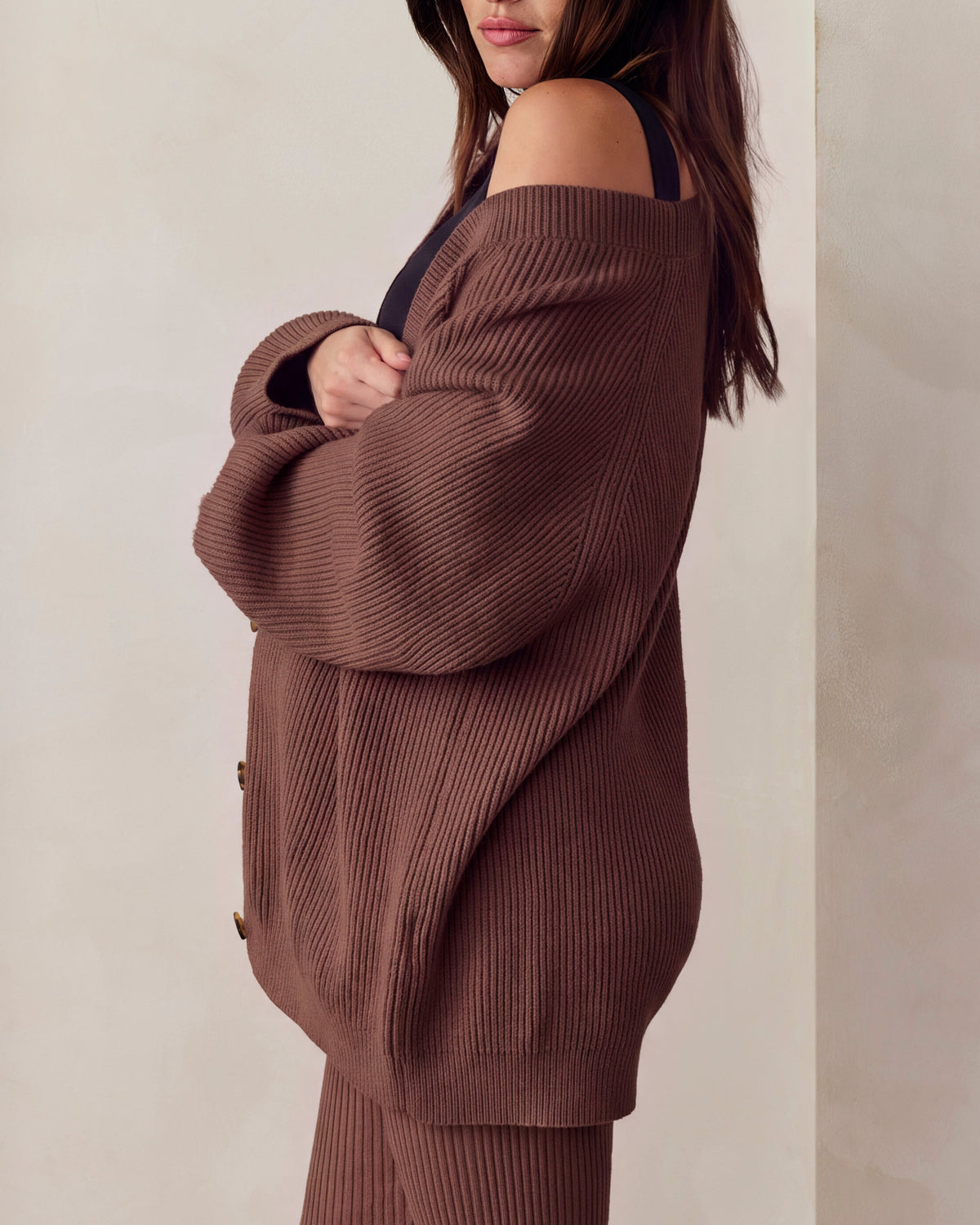 A pregnant woman wearing a matching brown ribbed knit cardigan and leggings set, standing against a neutral beige wall.