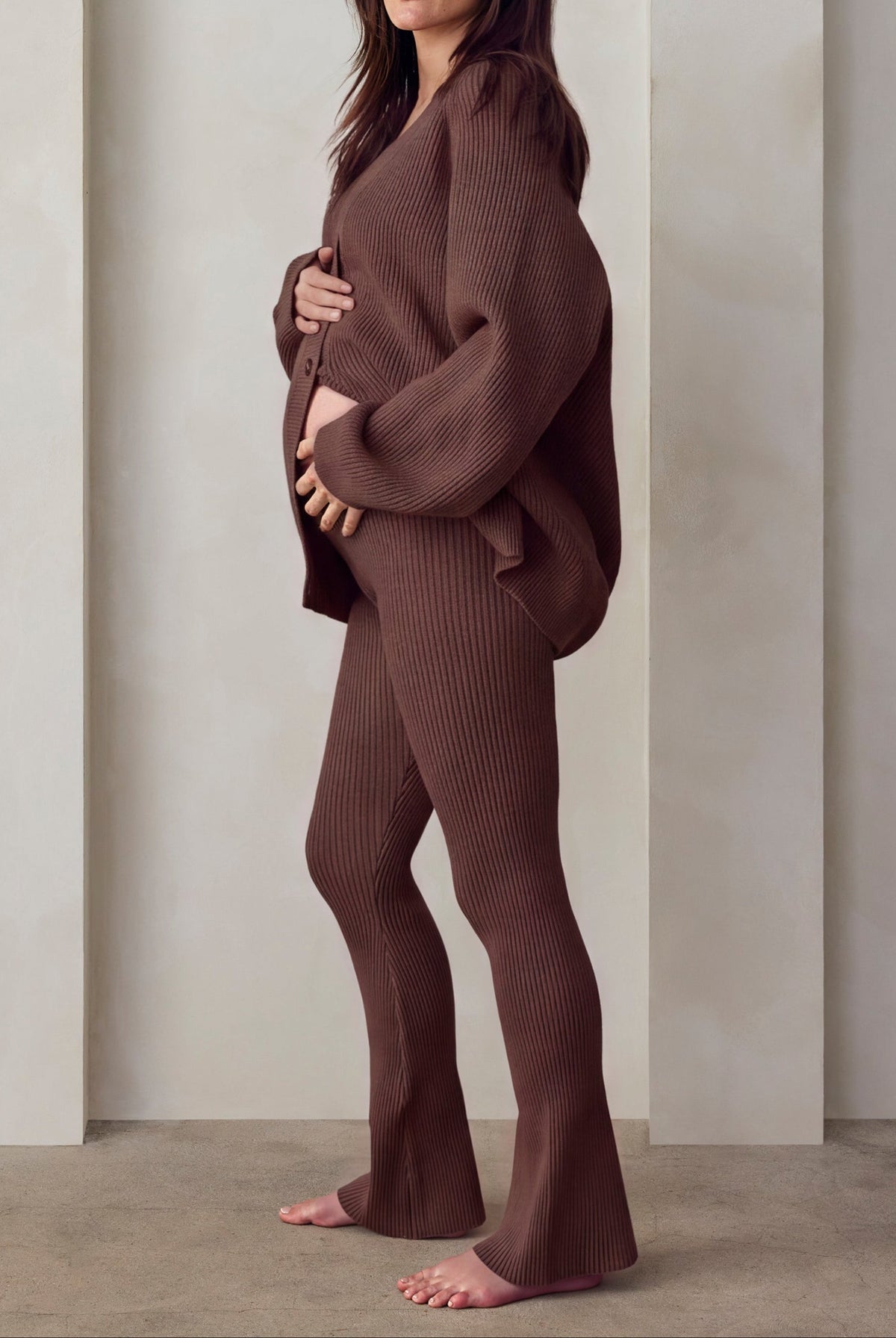 A woman stands barefoot in a minimalist studio setting with three arched recesses on a cream-colored wall. She is wearing a matching ribbed brown lounge set cyle top and flared pants.