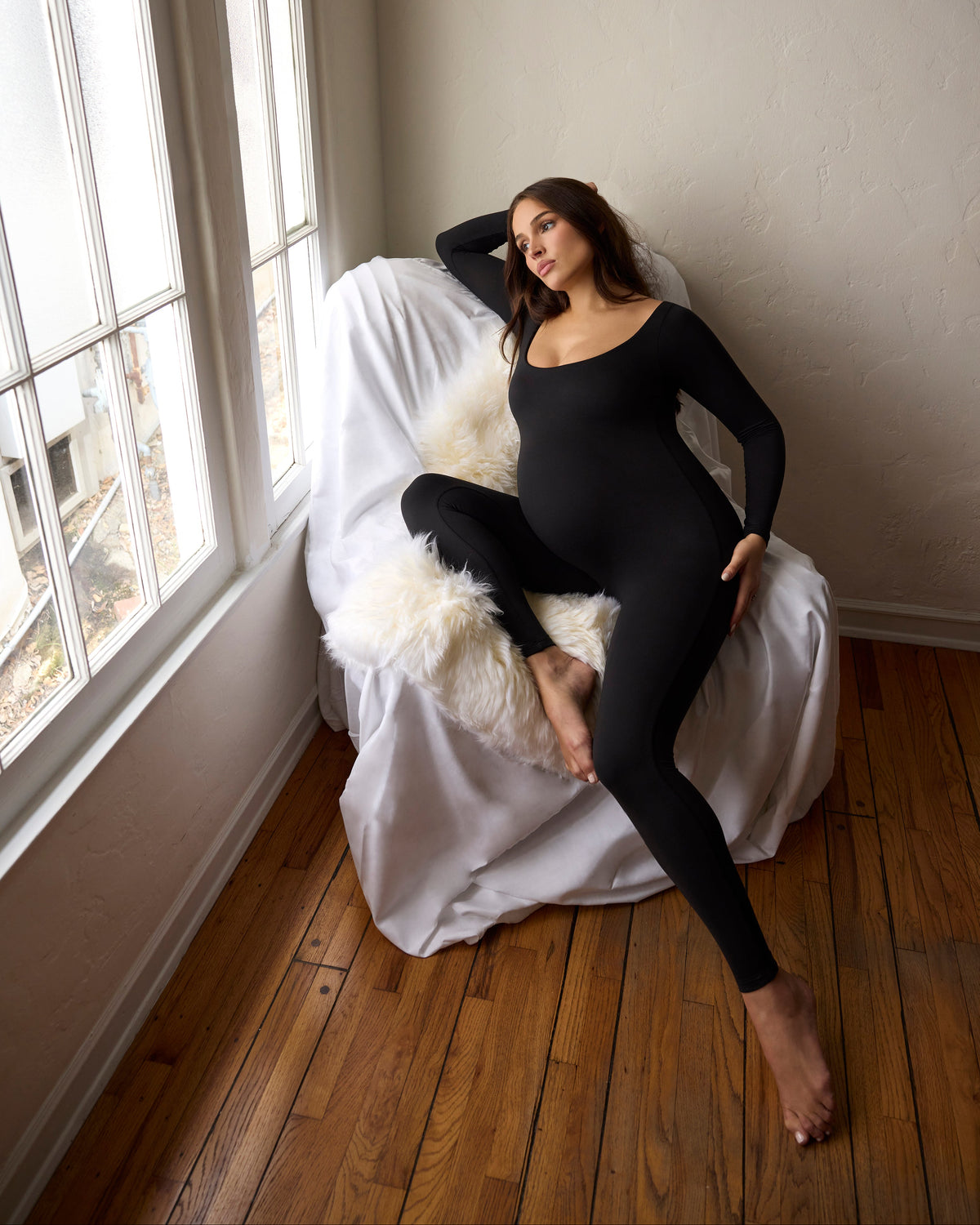 A pregnant woman wearing a black long-sleeve maternity jumpsuit sits on a white draped armchair with a fluffy white throw pillow in a room with hardwood floors and a large window.
