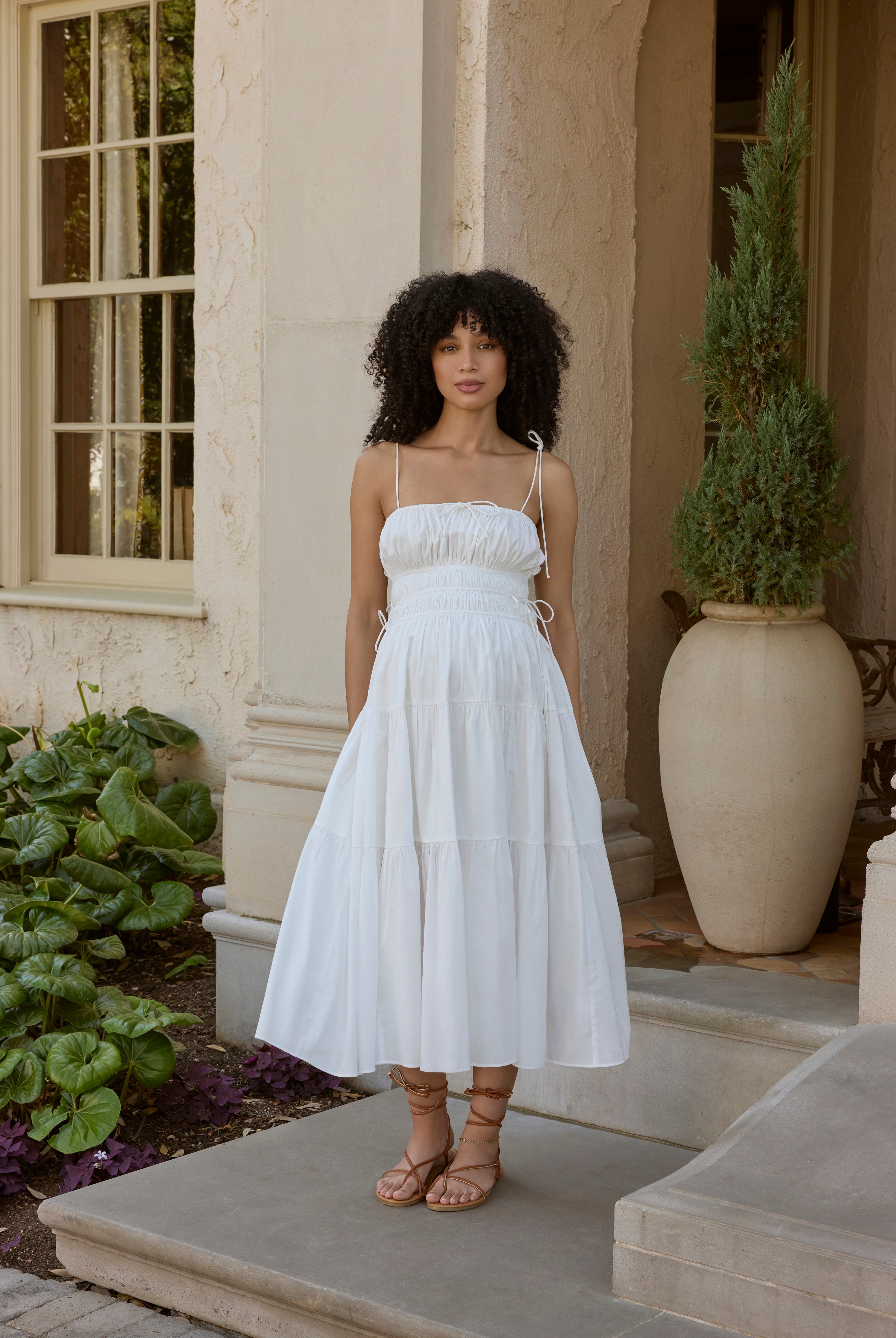 Woman in a white dress standing on a patio with plants and a building in the background