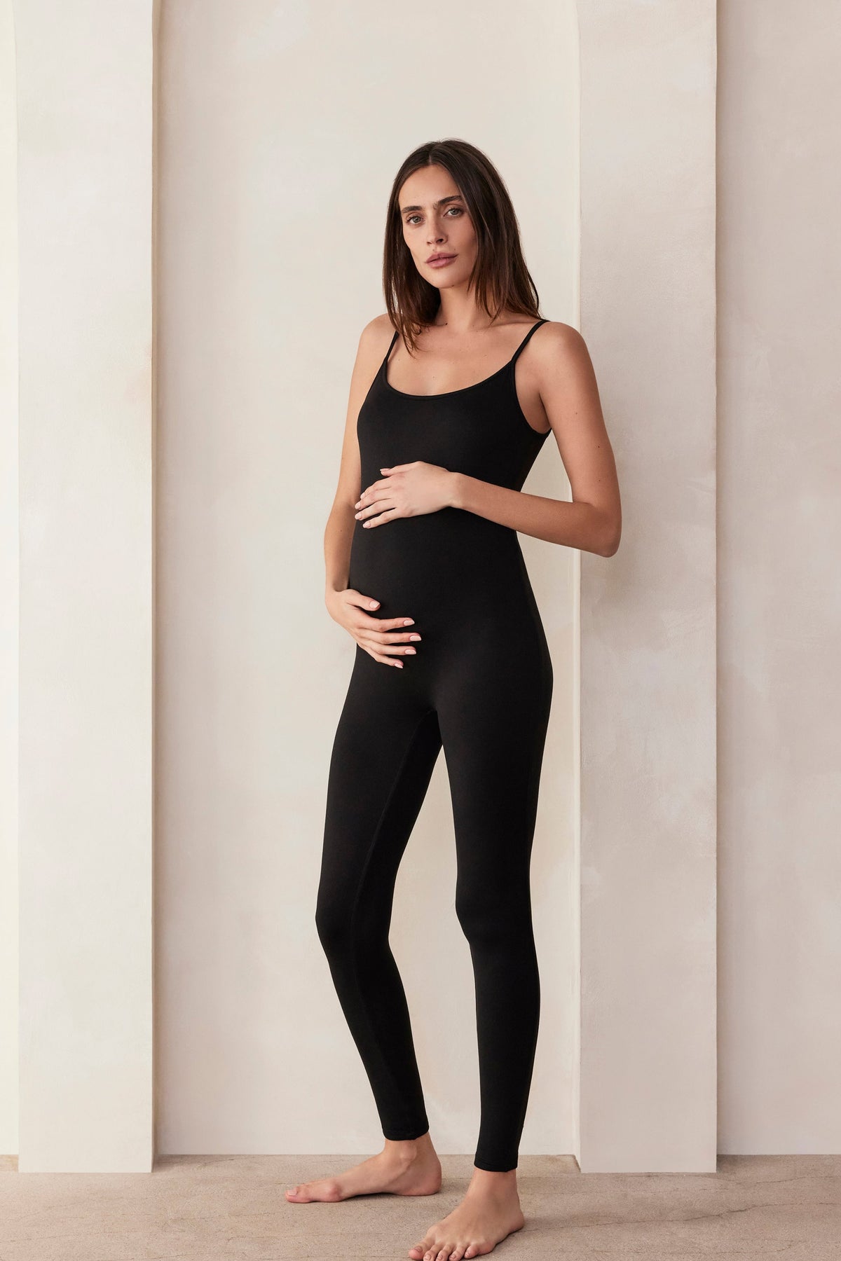 Woman wearing a Bumpsuit black sleeveless jumpsuit called the Kate standing in a minimalistic setting.