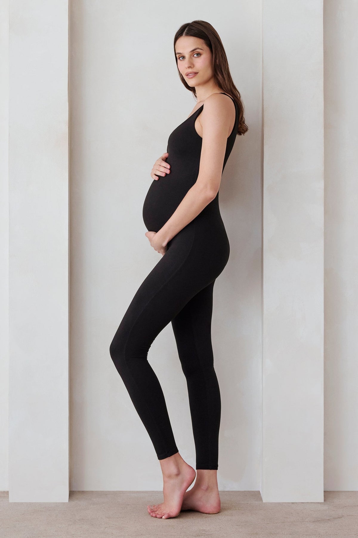 Side view of the black Cotton Lucy maternity unitard showcasing its full-body length fit on a pregnant model in a neutral studio