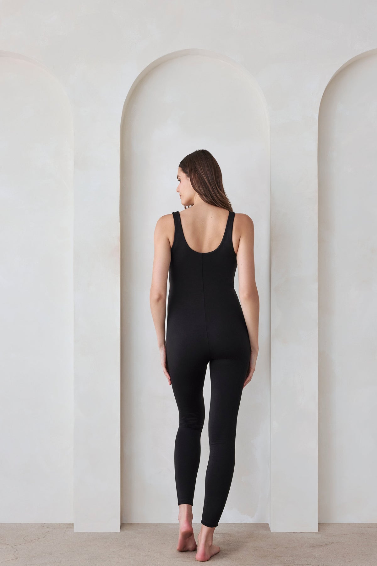 Back view of the black Cotton Lucy maternity unitard showcasing wide shoulder straps and full-body length on a model in a white arched room