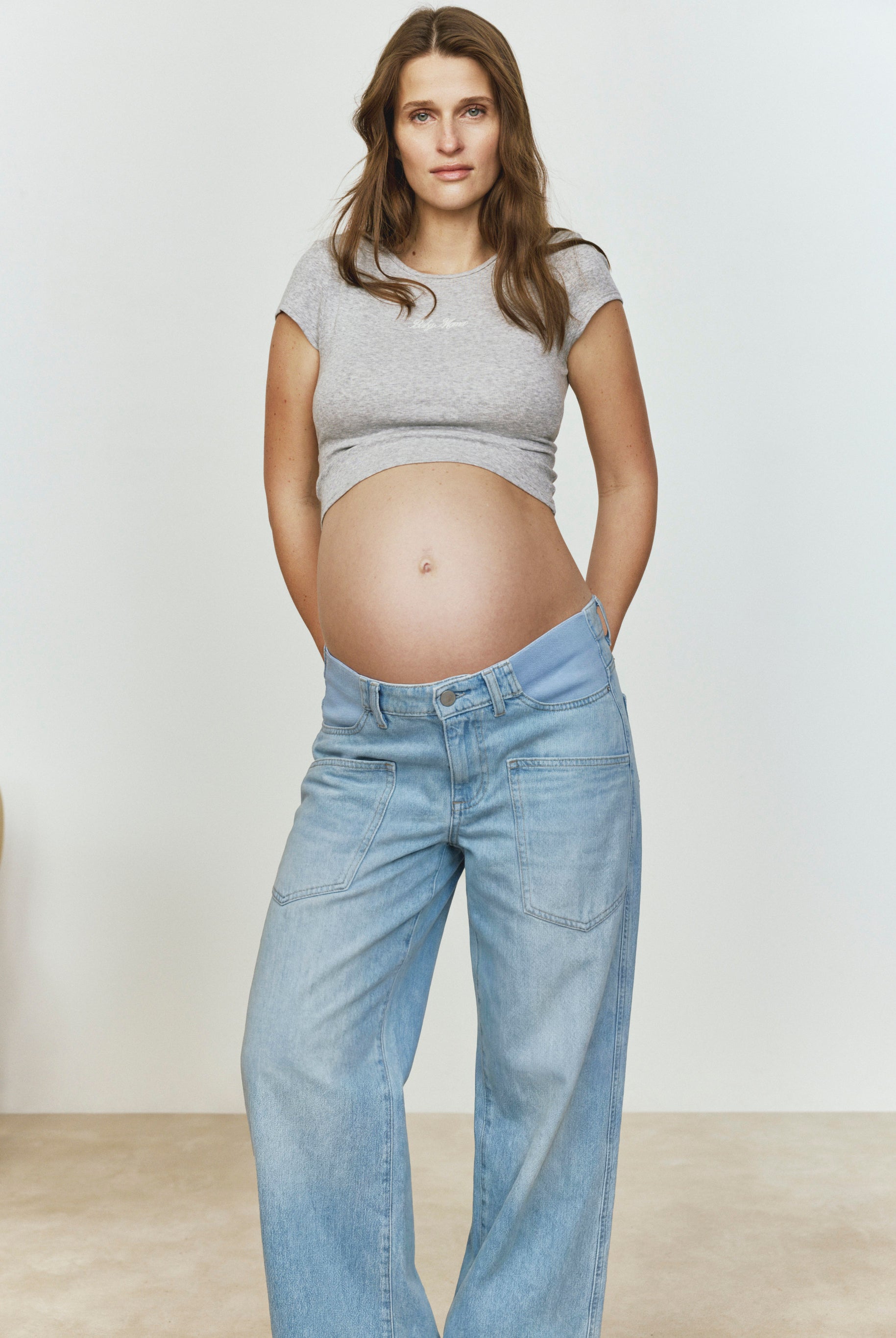 Pregnant woman wearing a heather gray baby mama tee and light blue jeans against a plain background
