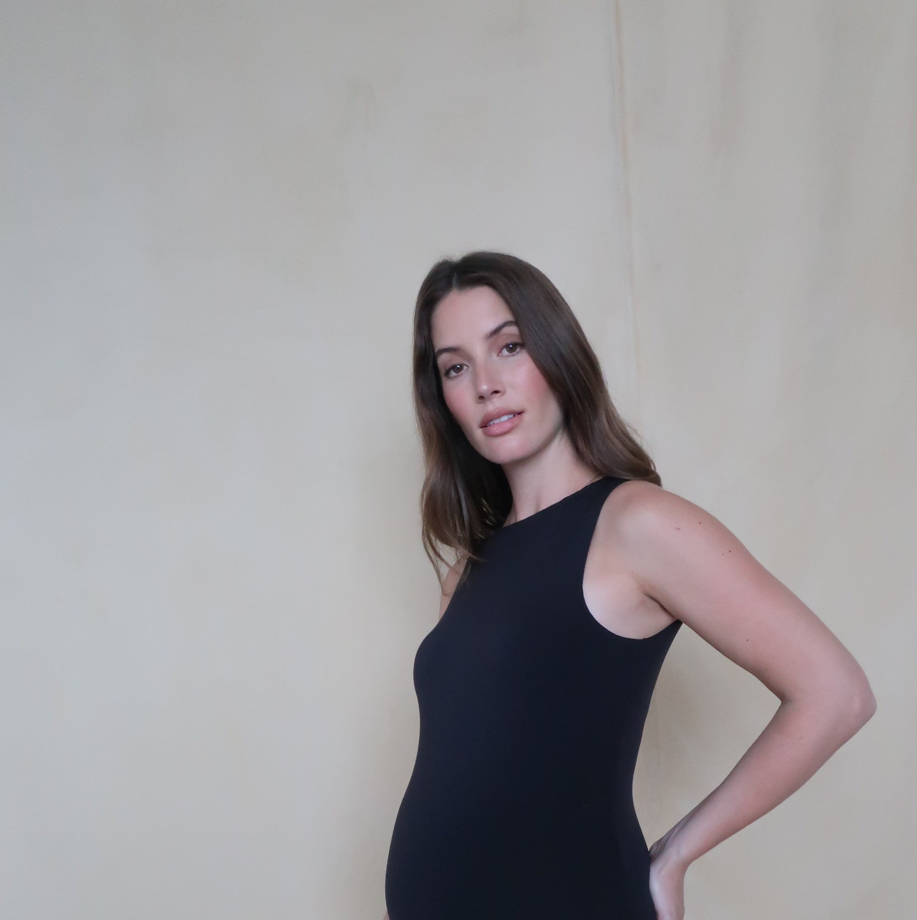 Pregnant woman wearing a black Bumpsuit bodysuit against a beige background