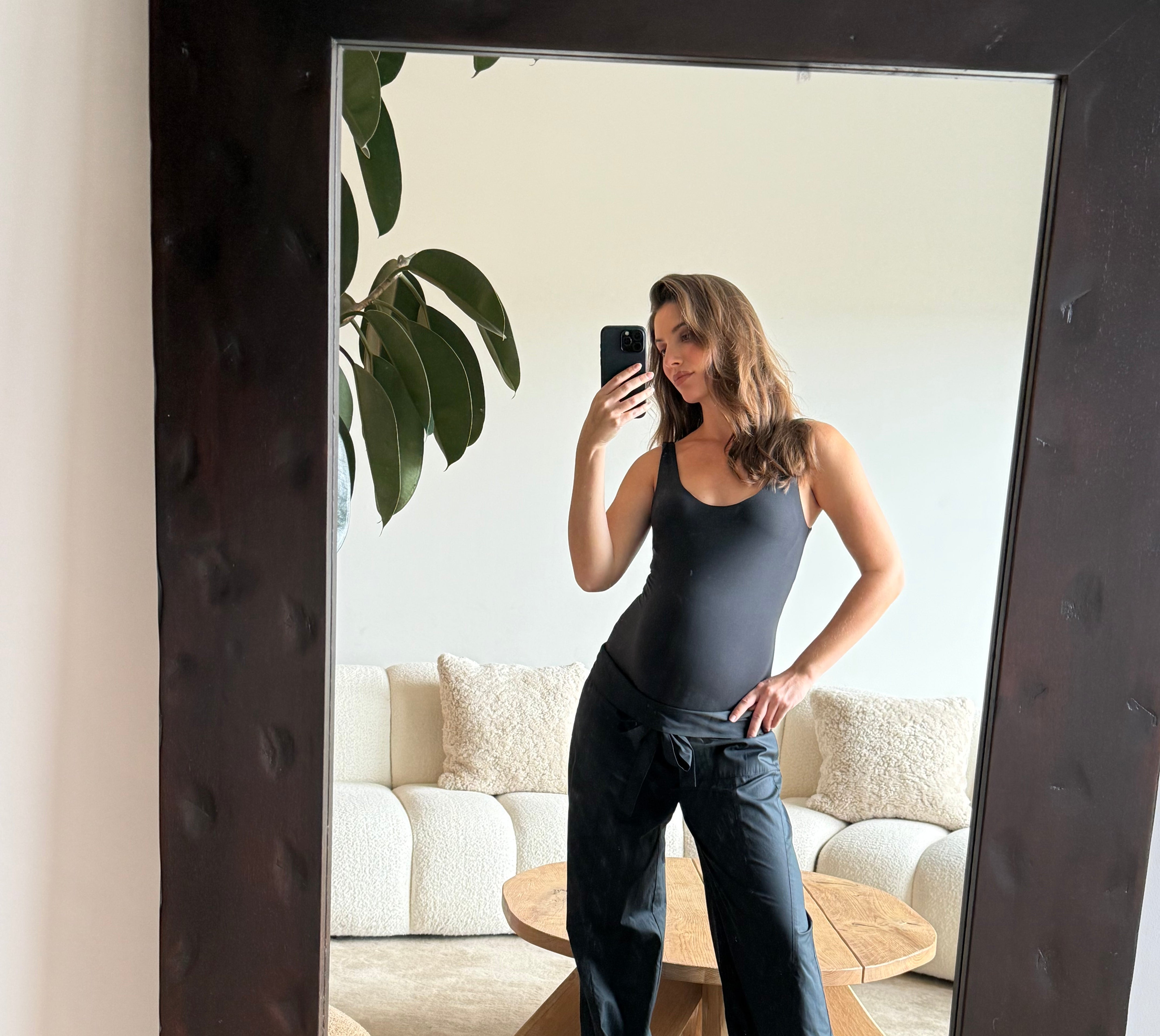 Pregnant women taking a mirror selfie in a modern living room wearing Bumpsuits tank bodysuit and jeans