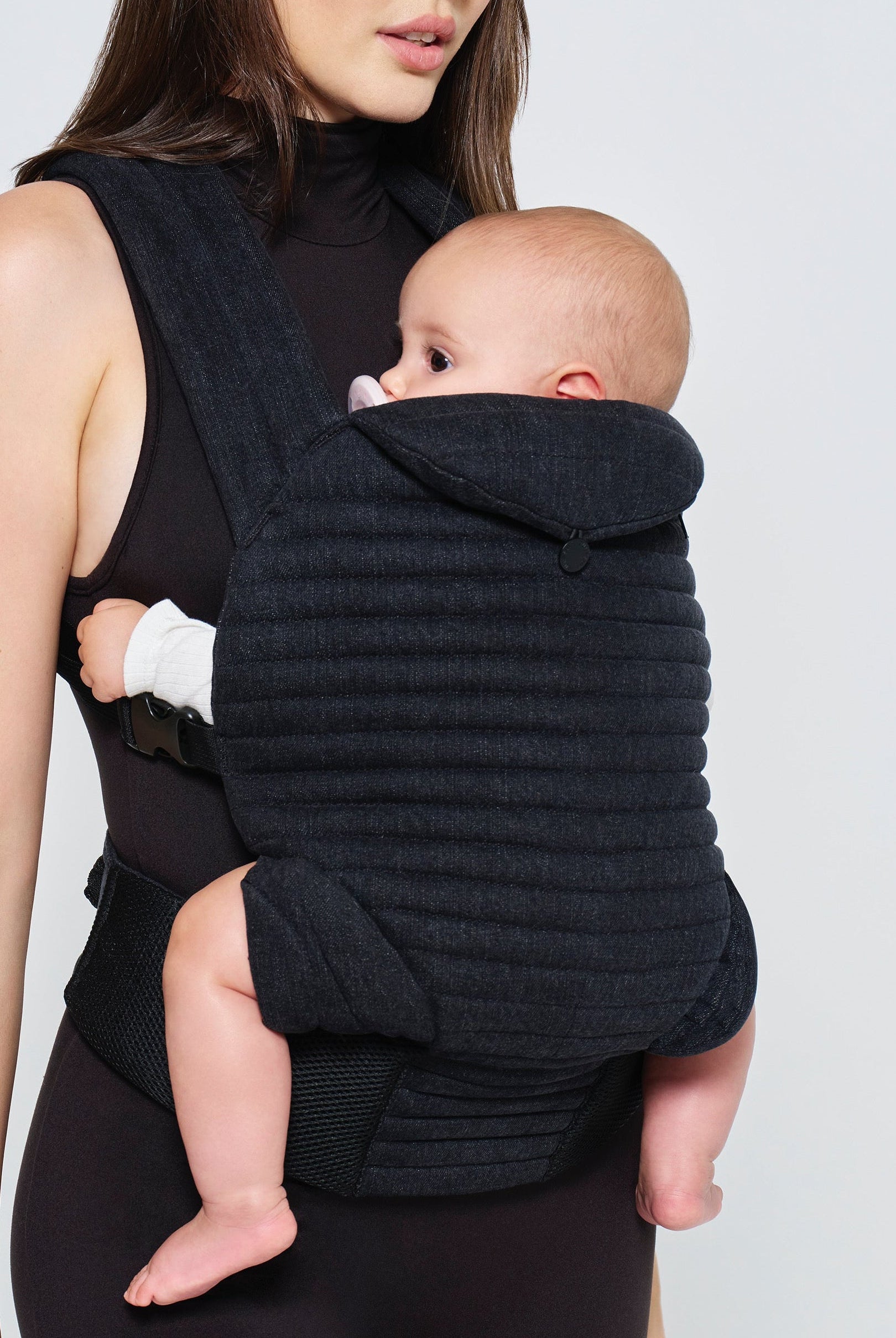 Woman holding a baby in a black denim baby carrier by Bumpsuit against a white background