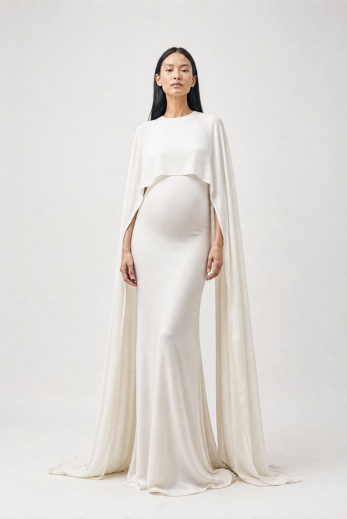 A pregnant model wearing a floor-length ivory maternity gown with a draped cape overlay, standing against a light gray studio background.
