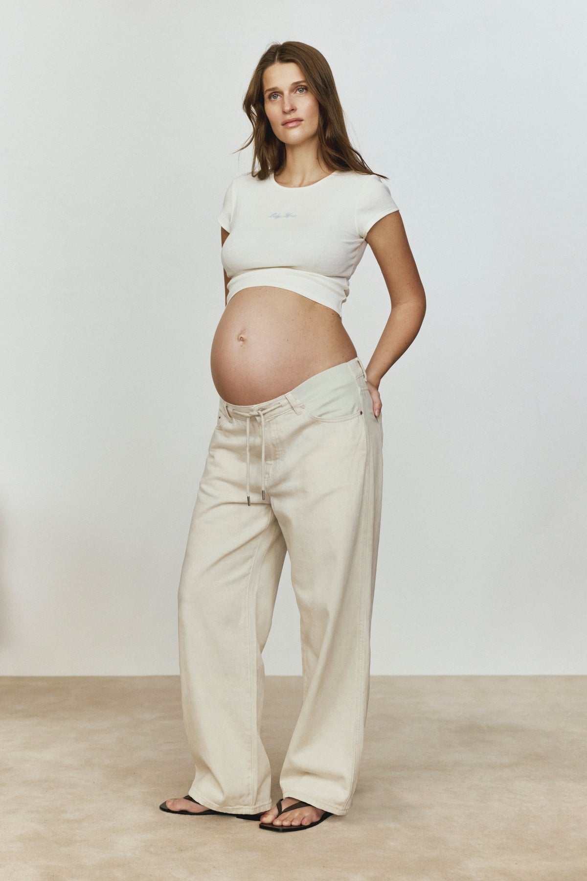 Pregnant woman wearing a white baby mama tee and beige pants against a plain background