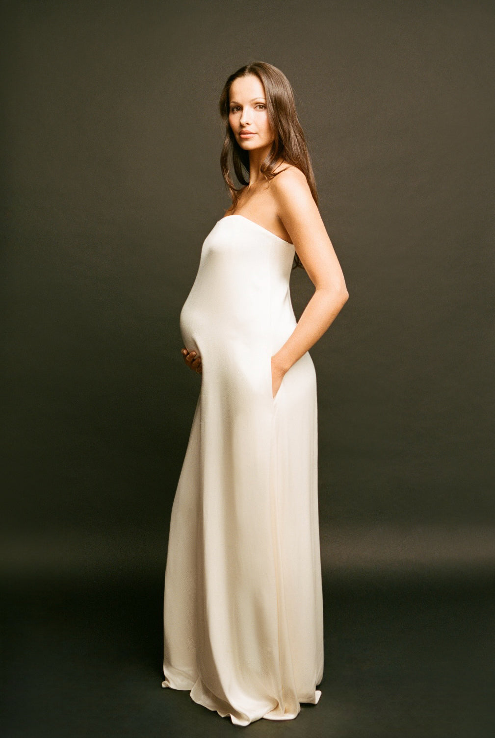 A pregnant woman wearing a Bumsuit strapless, floor-length cream satin gown stands against a dark studio background, holding her baby bump with one hand and resting the other in the side pocket of the dress.