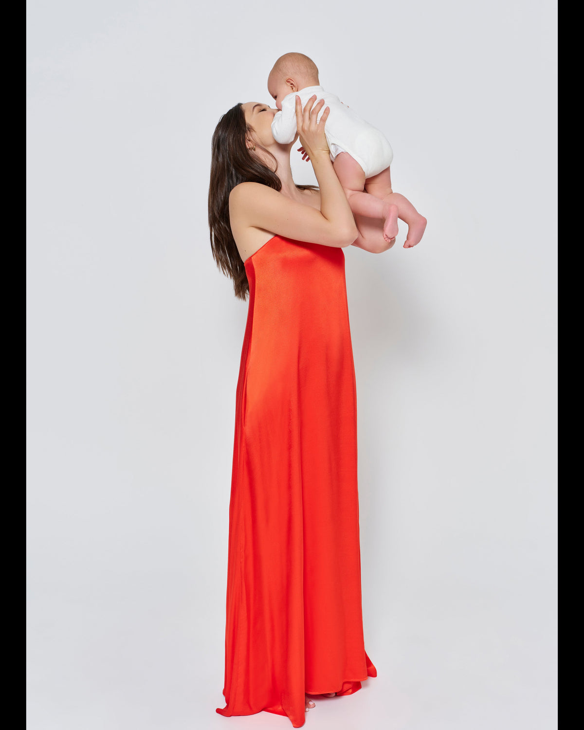 A woman wearing the Bumpsuit brushed satin strapless maxi maternity gown in red against a white background holding a baby in a white romper