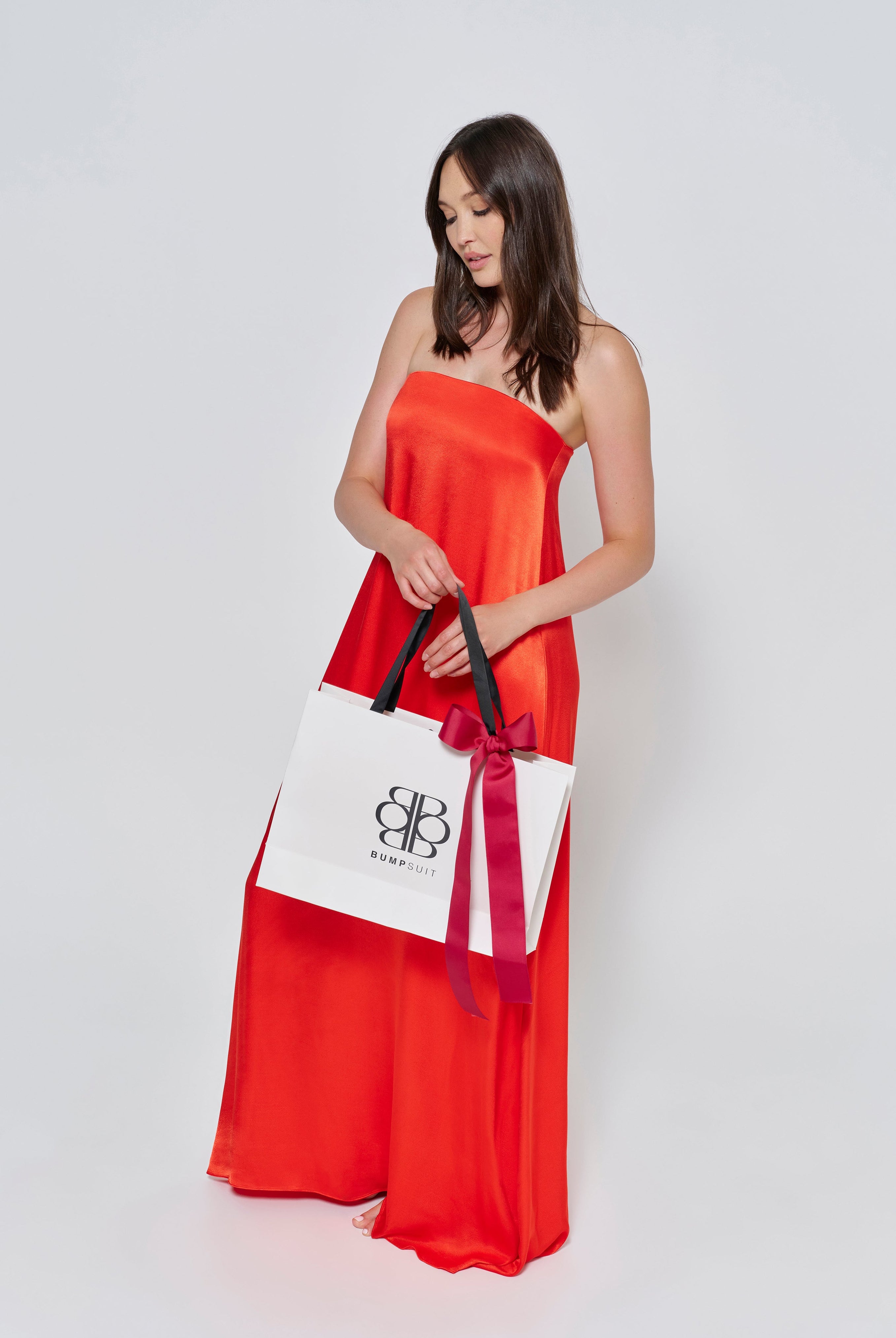A woman wearing the Bumpsuit brushed satin strapless maxi maternity gown in red against a white background holding a Bumpsuit gift bag
