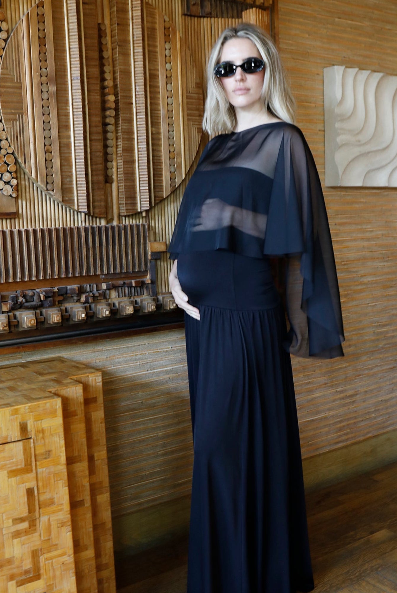 A woman wearing a long black maternity dress with a chiffon cape and sunglasses, standing in a rustic room with wood-paneled walls and wooden furniture.