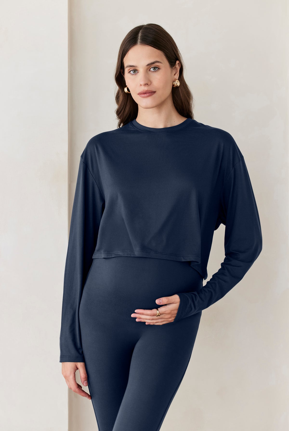 A woman standing against a beige feature wall wearing Bumpsuits Cloud long sleeve crop tee in navy