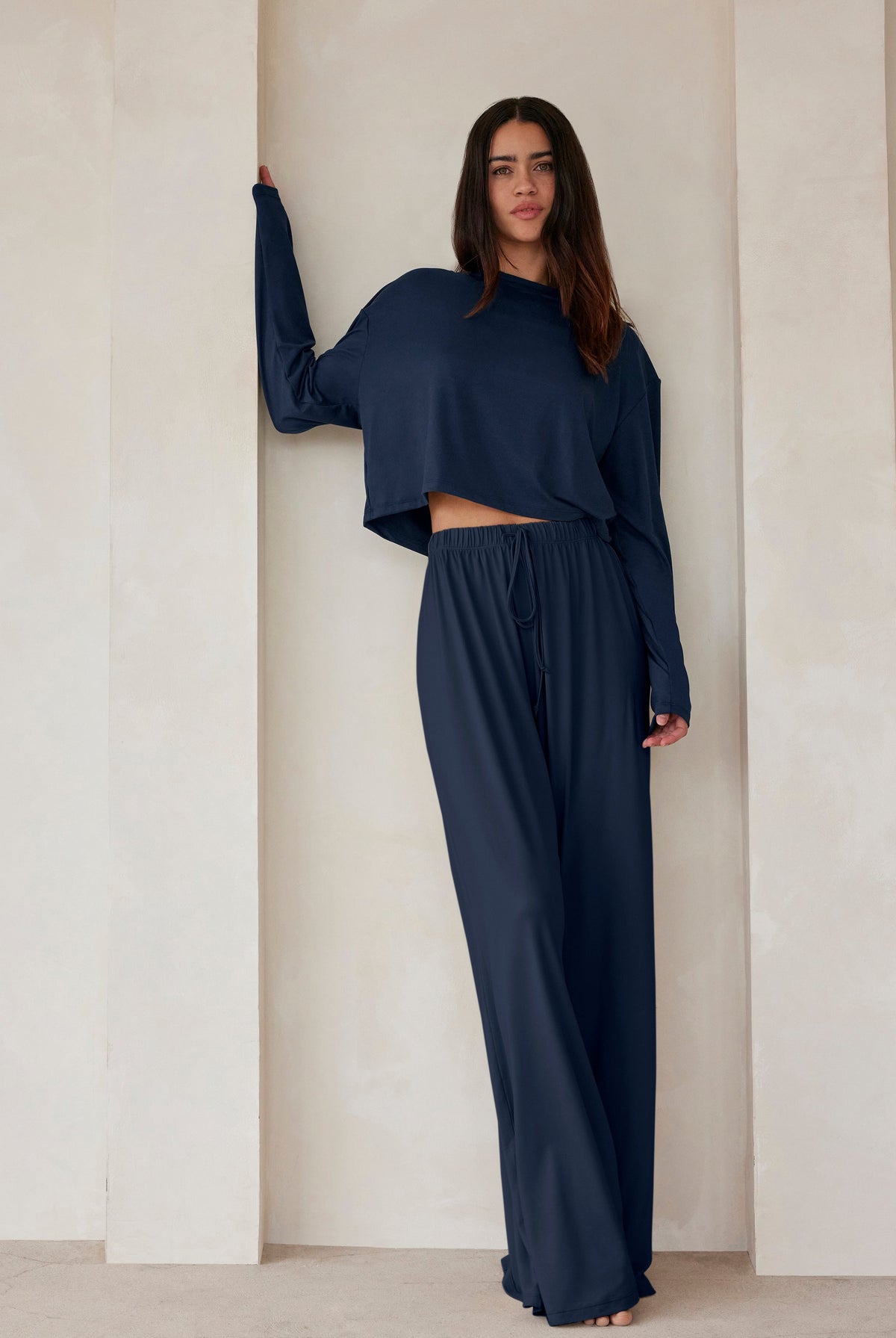 A woman standing against a beige feature wall wearing Bumpsuits Cloud lounge pants and a long sleeve crop tee in navy