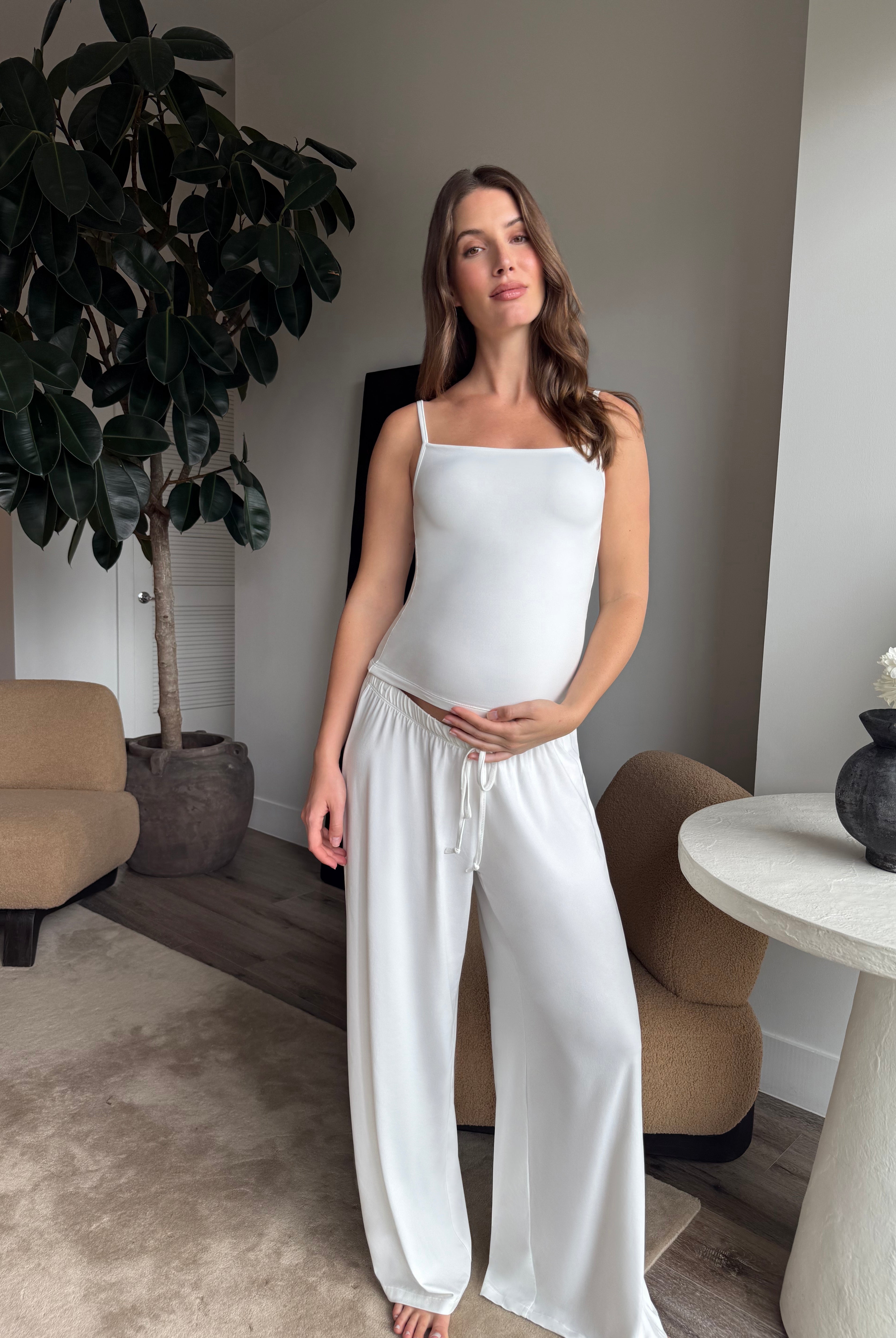 Pregnant woman wearing a Bumpsuit cloud ice pink tank top and pants in a living room.