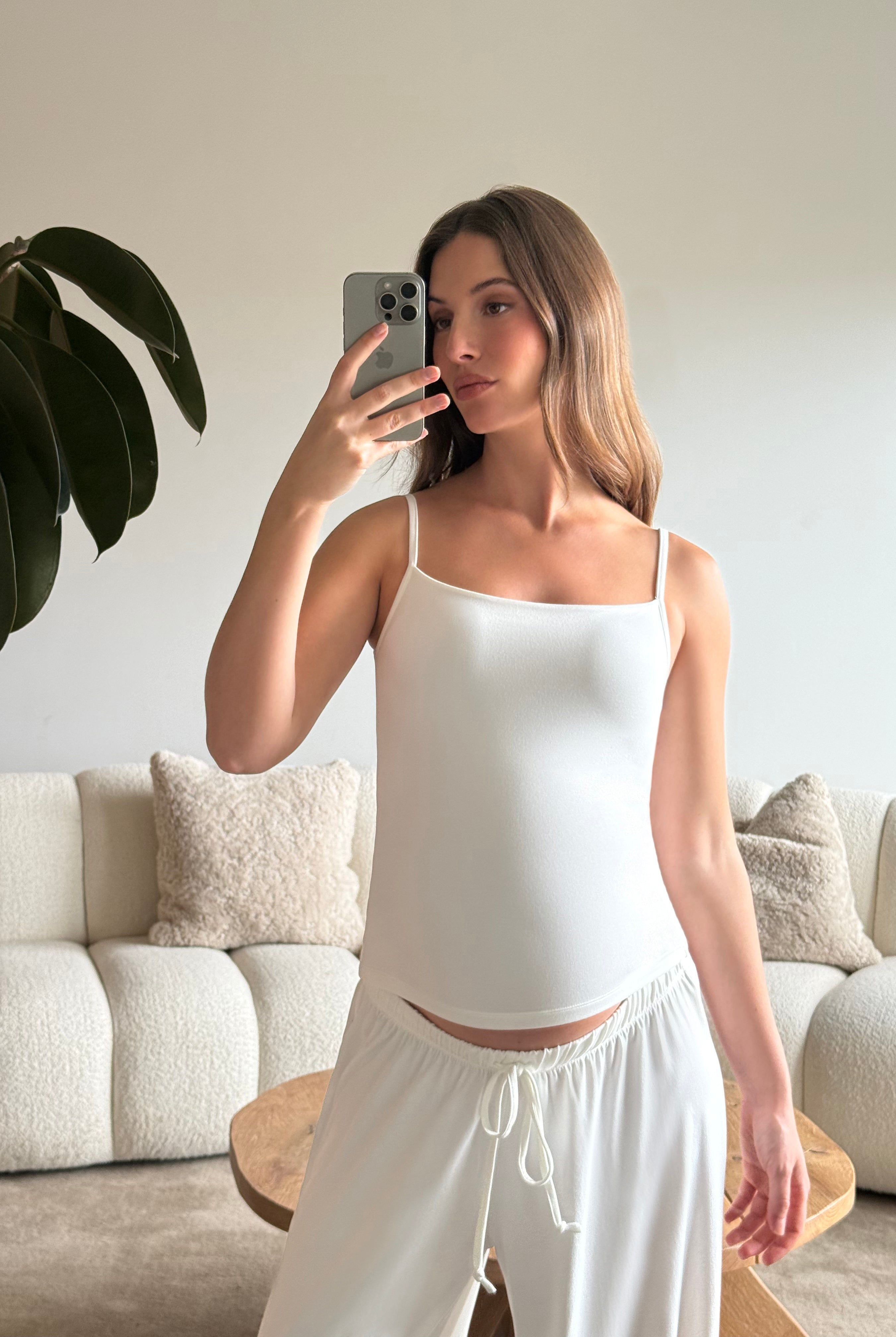 Pregnant woman taking a mirror selfie wearing a Bumpsuit cloud ivory tank top and pants in a living room.