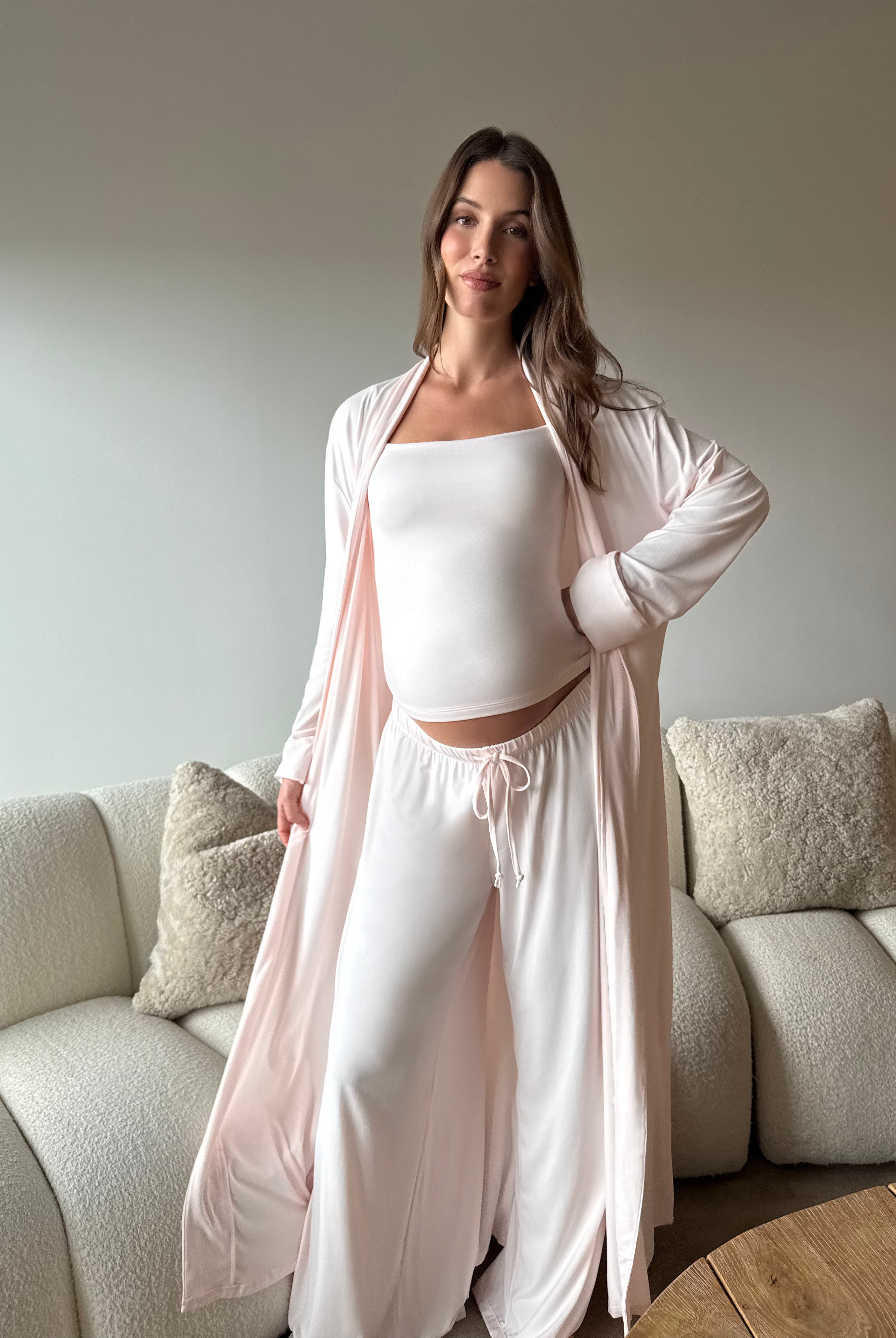Pregnant woman taking a mirror selfie wearing a Bumpsuit cloud ice pink tank top, robe and pants in a living room.