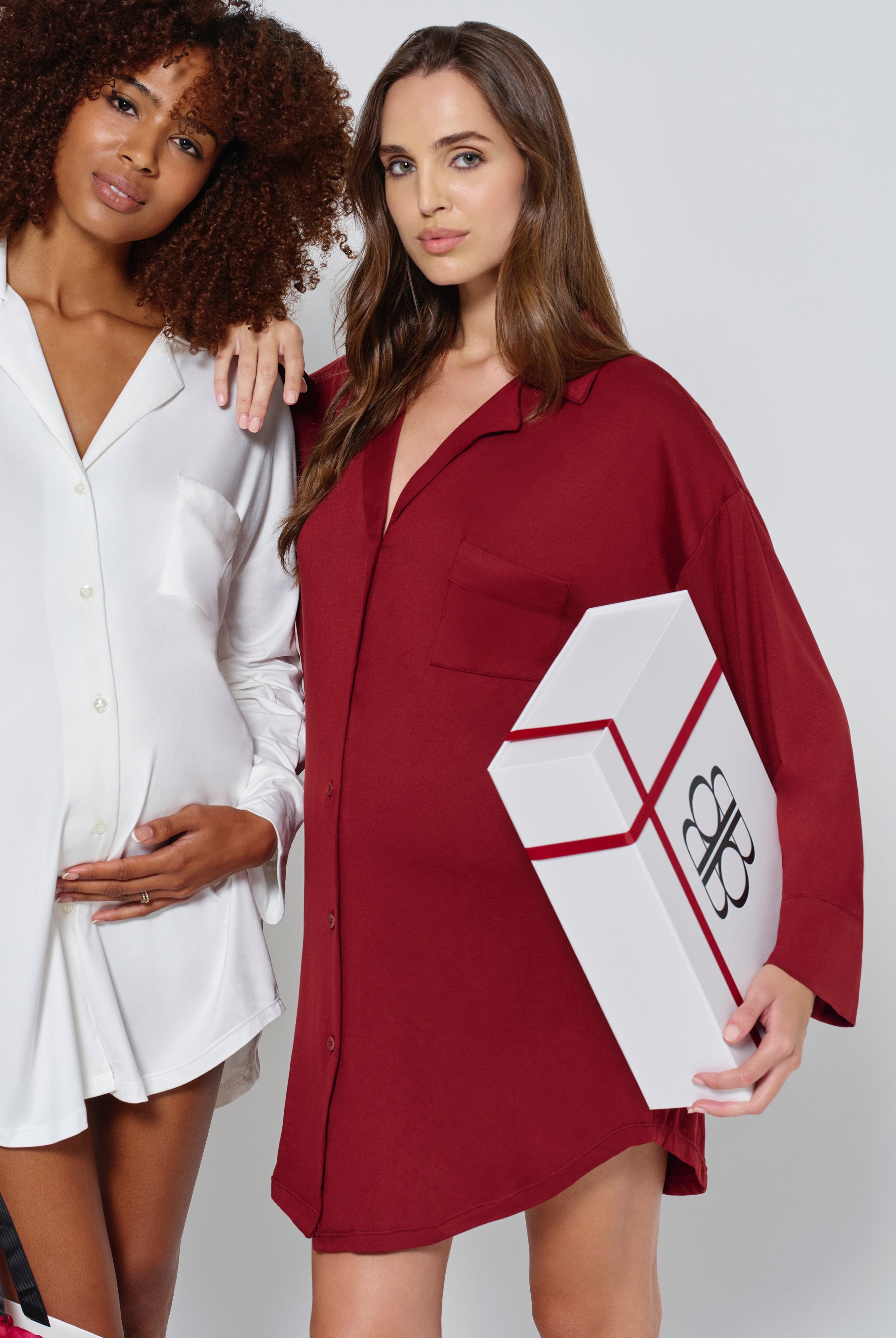 two pregnant women dressed in Bumpsuits Pajama dress, one in ivory and the other in burgundy, holding gift boxes on a white background