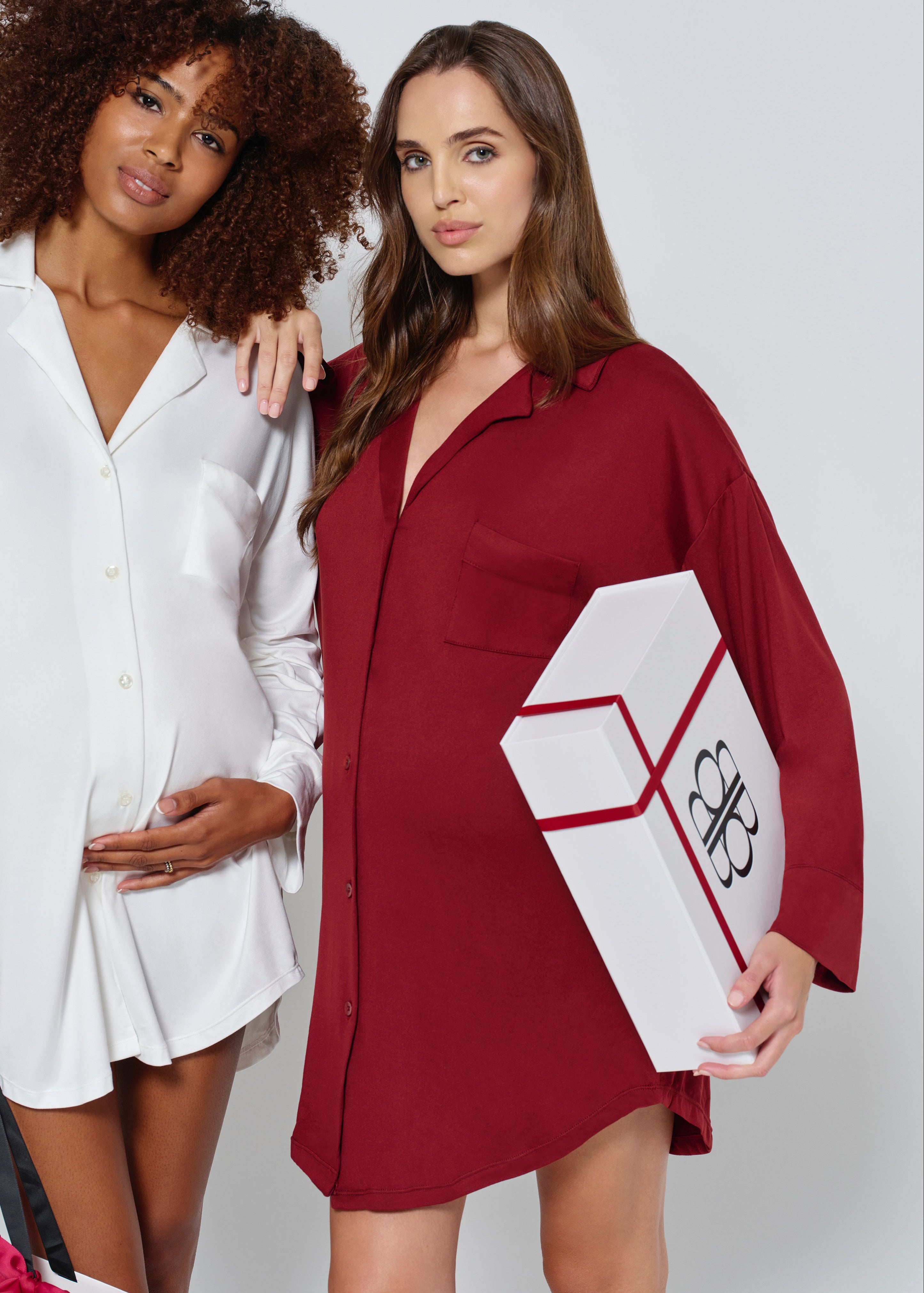 two pregnant women dressed in Bumpsuits Pajama dress, one in ivory and the other in burgundy, holding gift boxes on a white background
