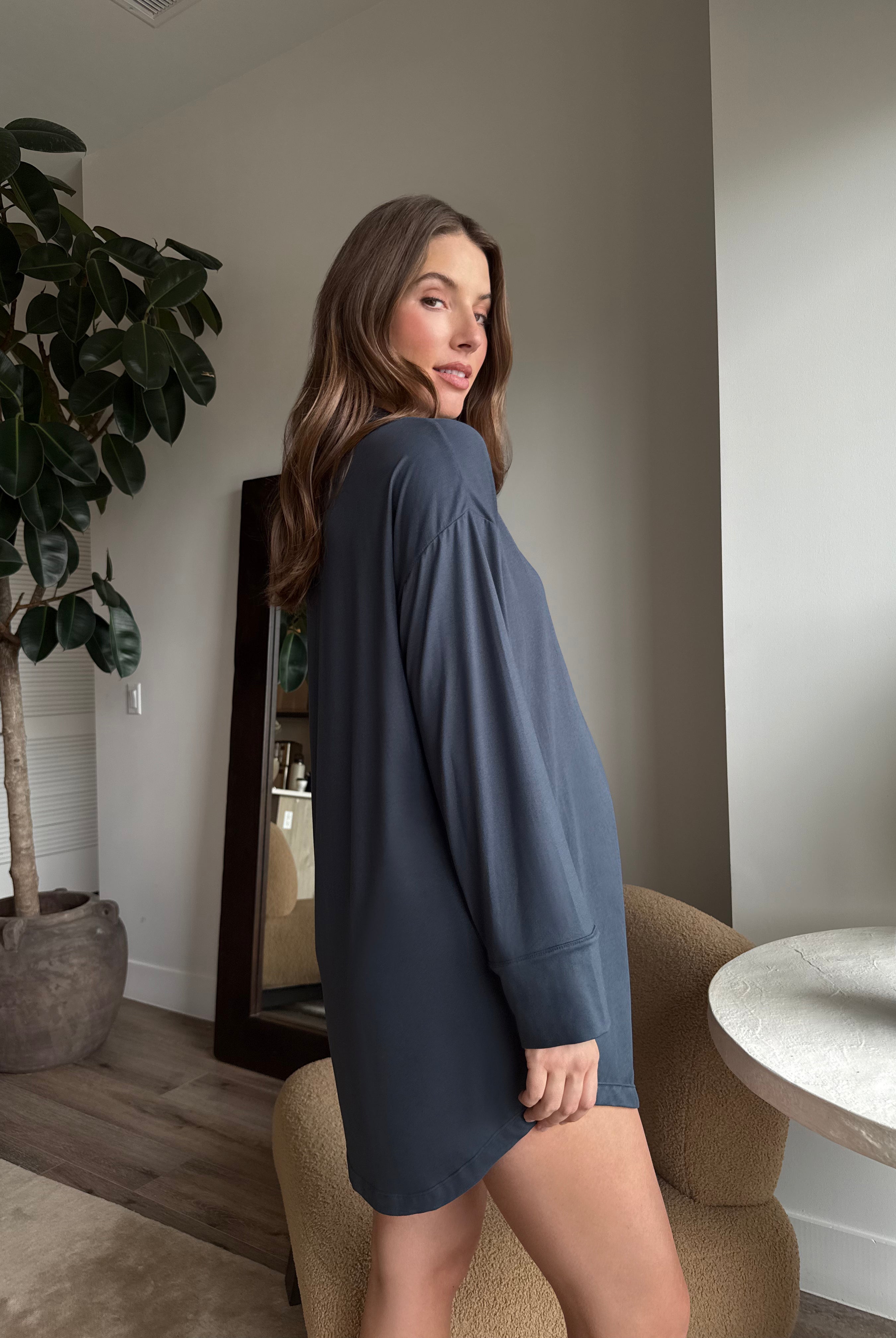 Pregnant woman wearing a Bumpsuit Cloud cement blue pyjama dress standing in a room with a plant and table.
