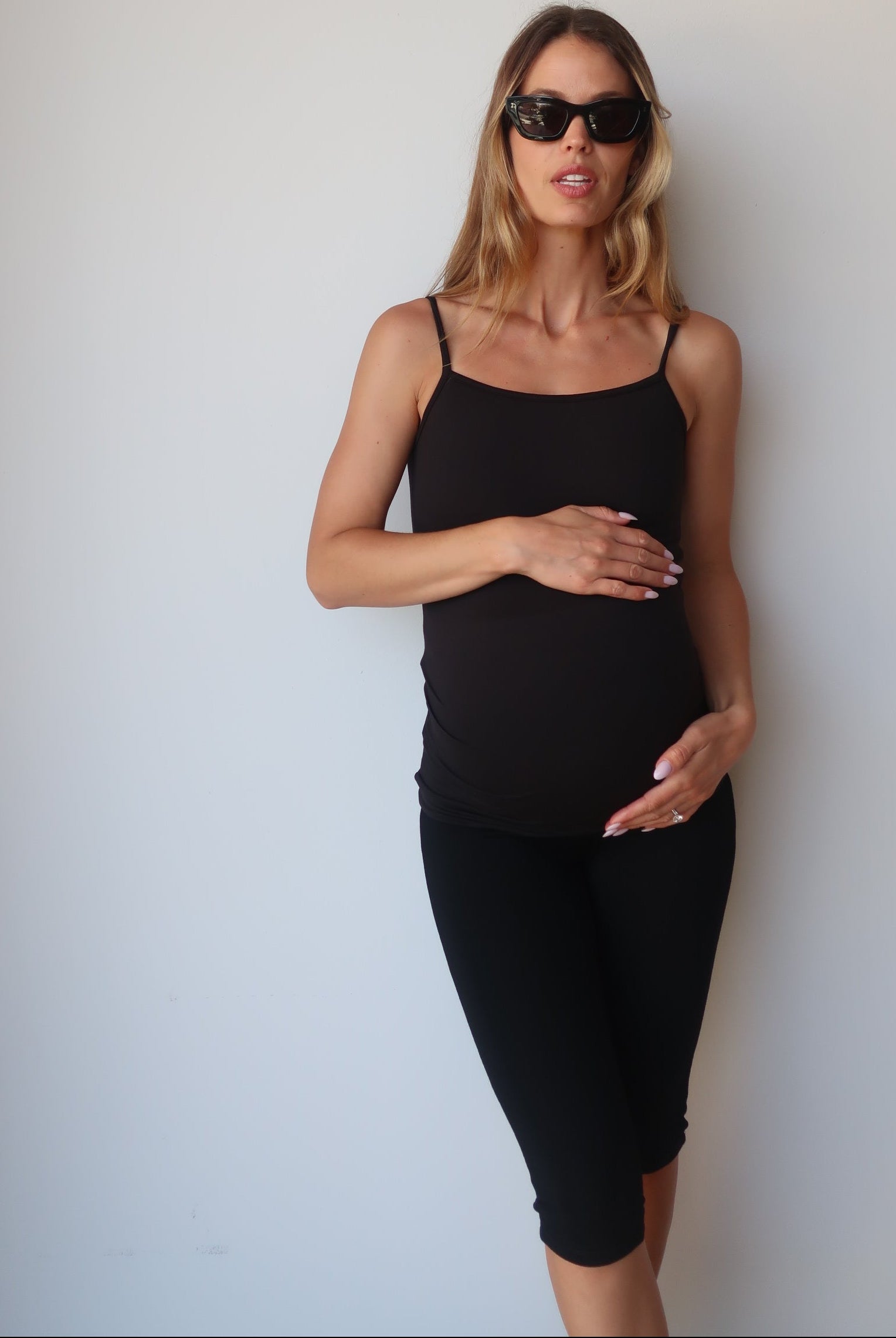 Pregnant woman wearing a black Bumpsuit camisole tank top and capri leggings against a white wall