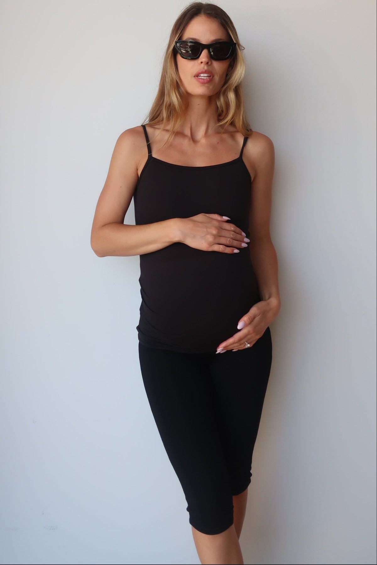 Pregnant woman wearing a black Bumpsuit camisole tank top and capri leggings against a white wall