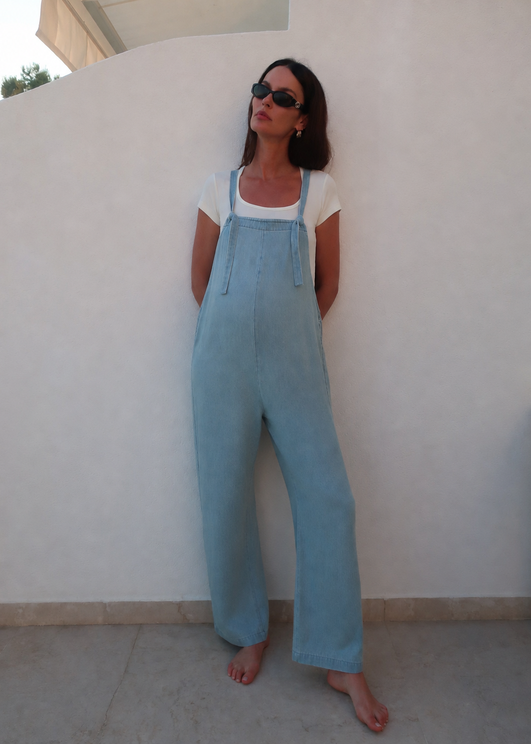 A woman wearing a light blue denim overall jumpsuit over a white t-shirt, standing barefoot against a white wall.