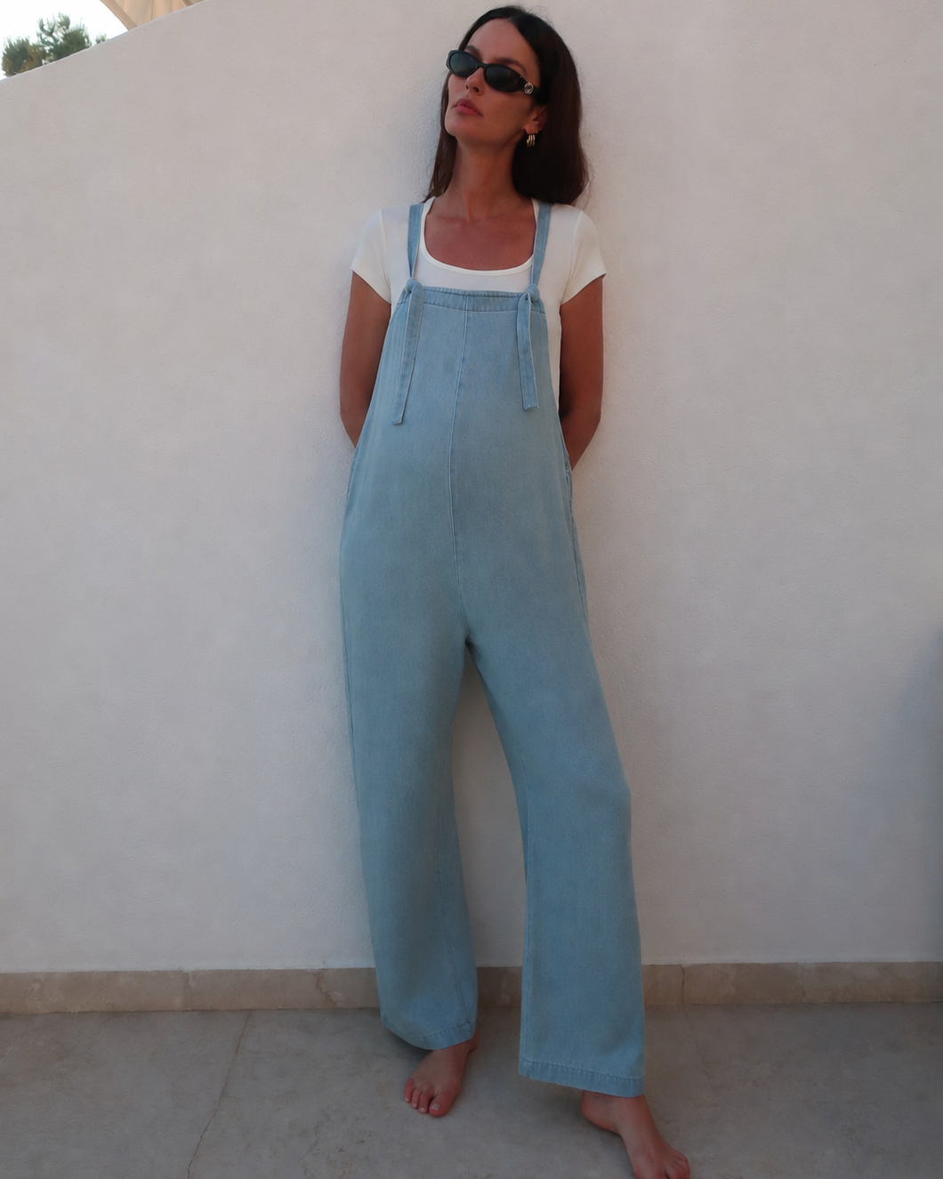 A woman wearing a light blue denim overall jumpsuit over a white t-shirt, standing barefoot against a white wall.