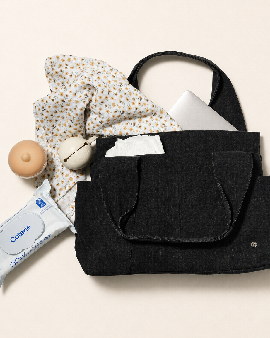 A black denim fabric diaper bag shown in a styled flat-lay with a laptop, patterned scarf, milk bottle, and wipes packet around it on a warm off-white background.
