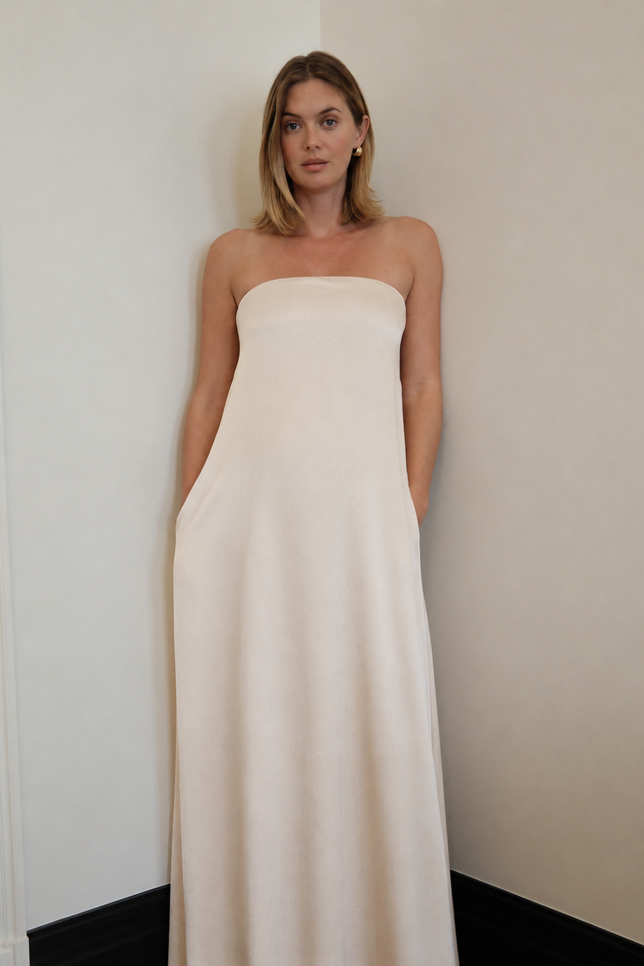A pregnant woman standing with her hands in her pockets, in the corner of a room wearing a champagne coloured, brushed satin strapless maternity gown.