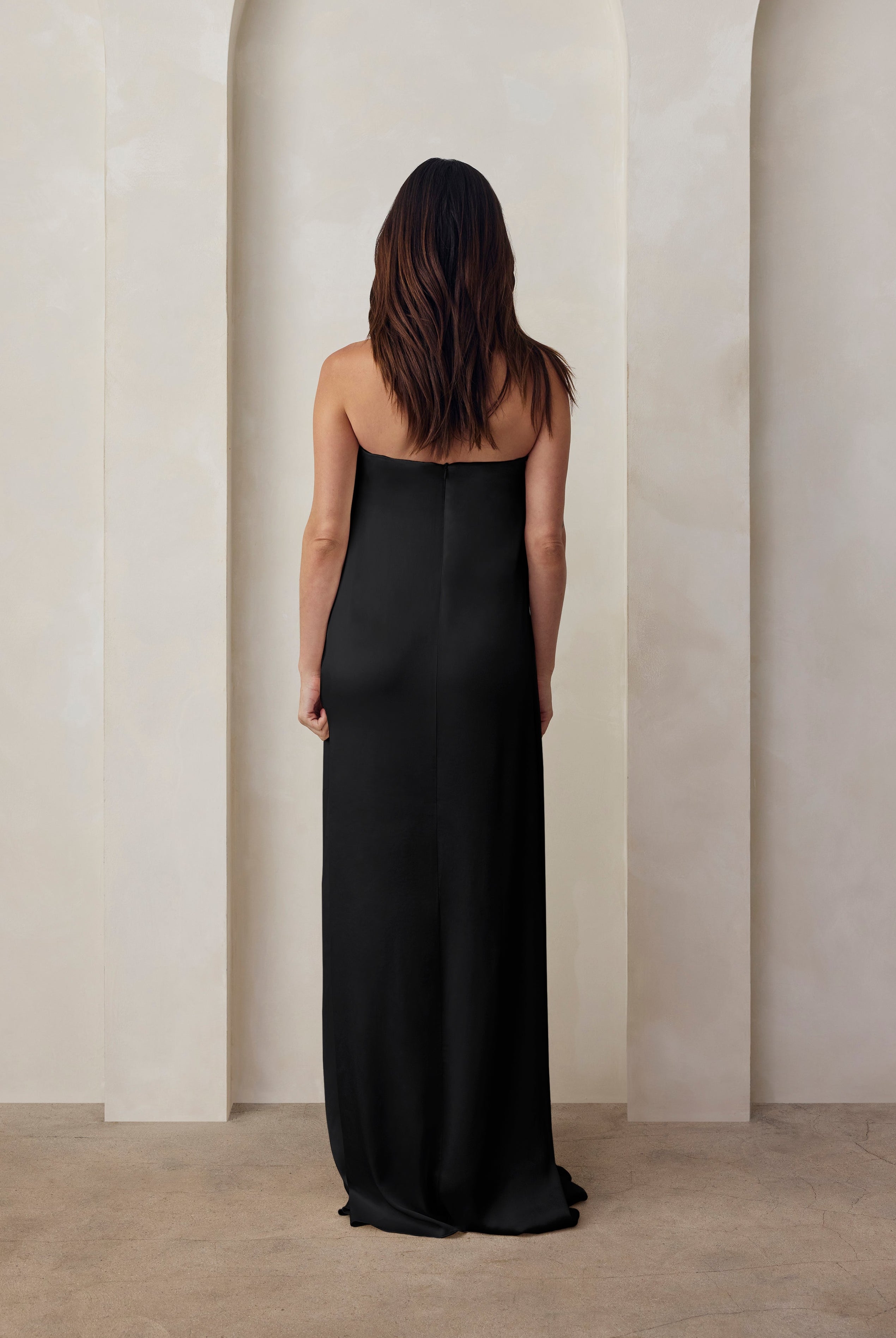 Back view of a pregnant woman wearing the Bumpsuit brushed satin strapless maxi maternity gown in black against a beige feature wall