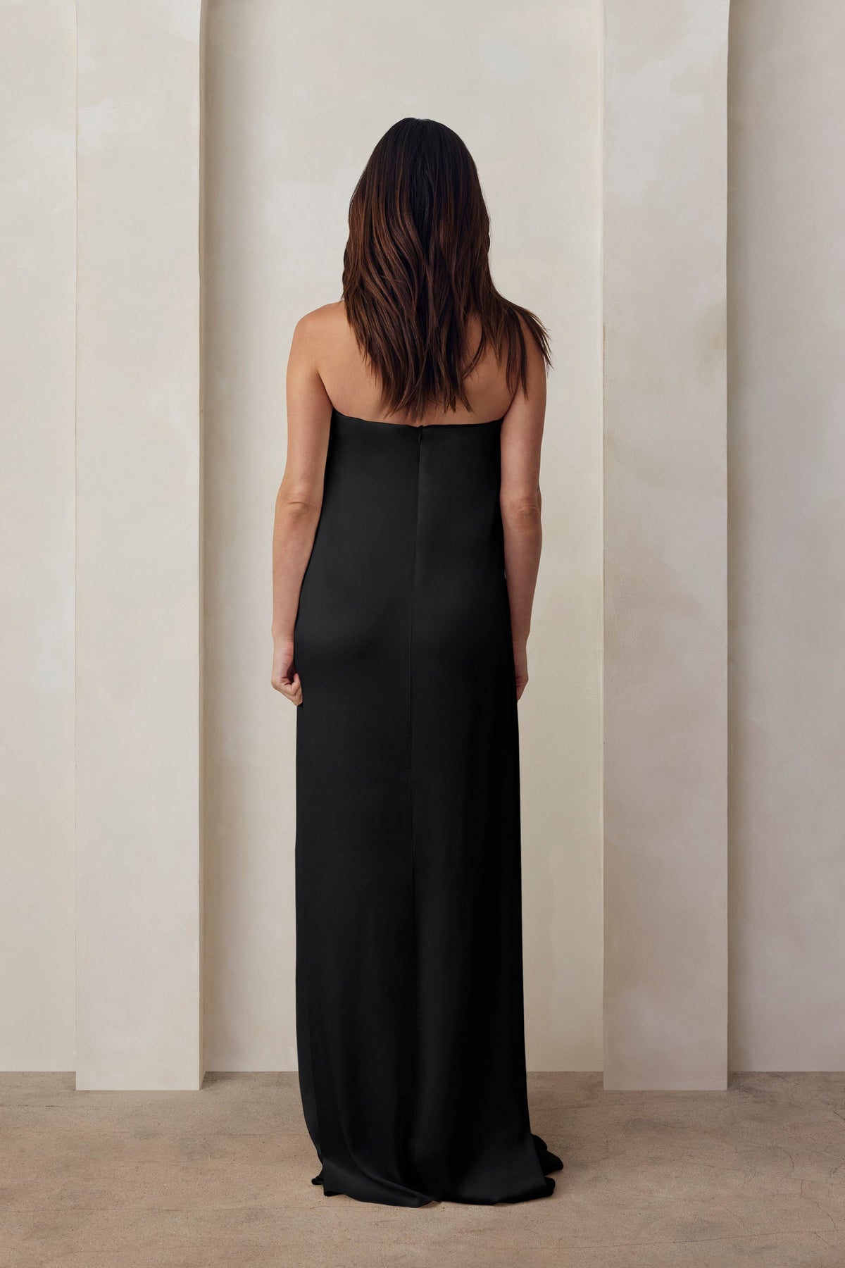 Back view of a pregnant woman wearing the Bumpsuit brushed satin strapless maxi maternity gown in black against a beige feature wall
