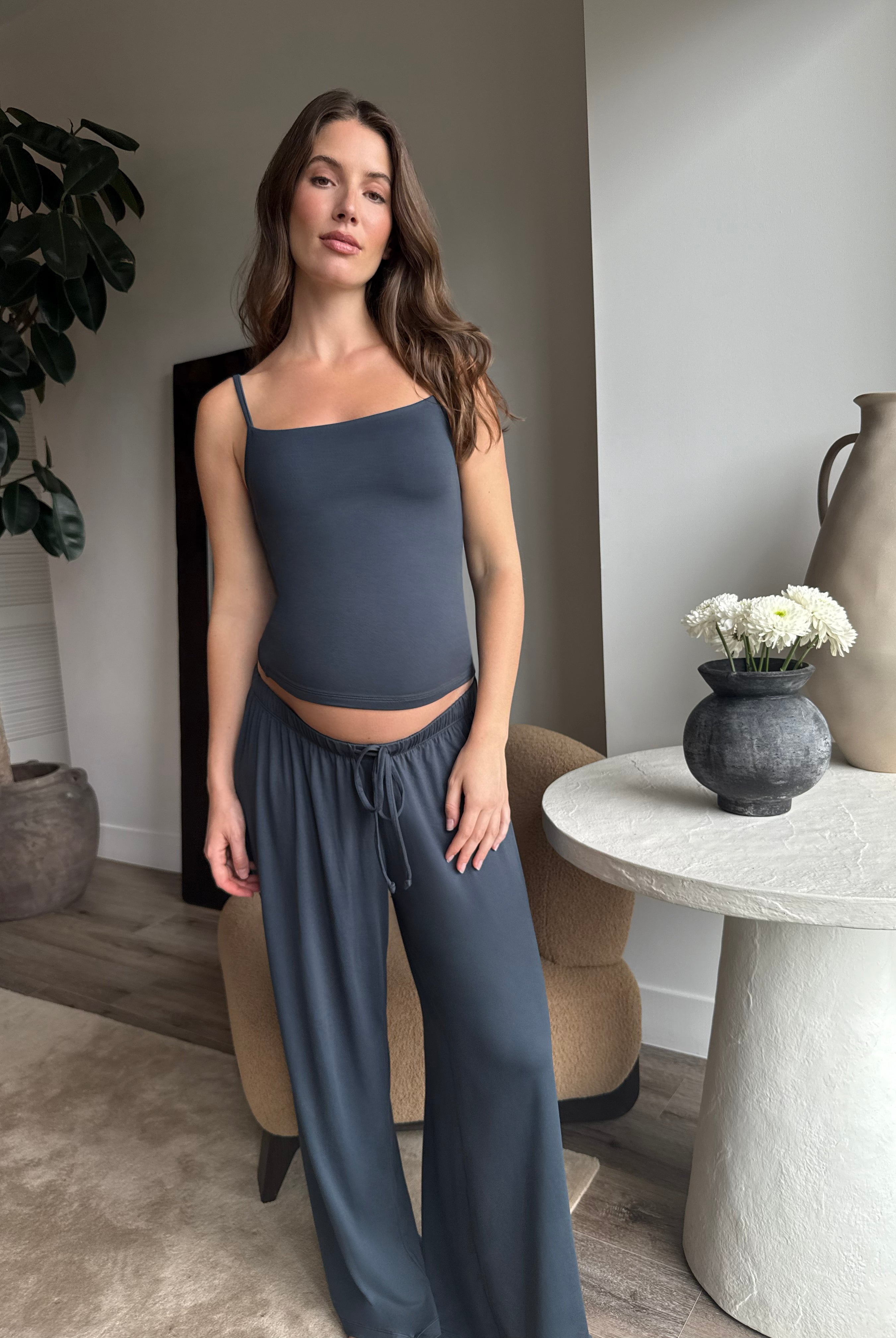 Pregnant woman wearing a Bumpsuit cloud cement blue tank top and pants in a living room.